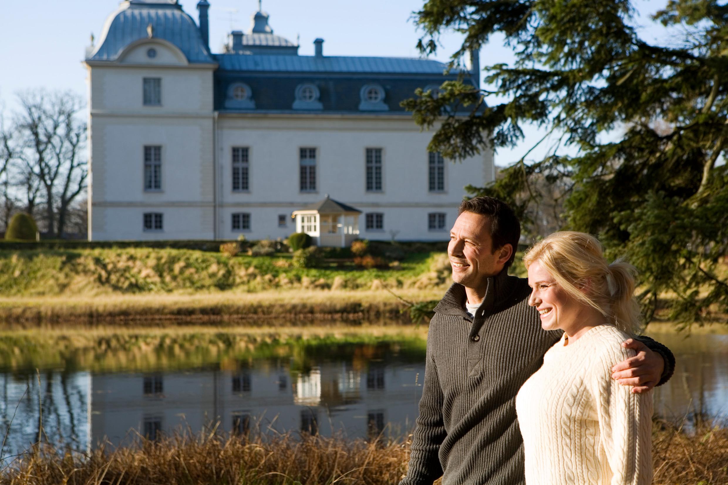 Un couple se promène main dans la main au bord de l'eau, avec le château viticole de Kronovall en arrière-plan.