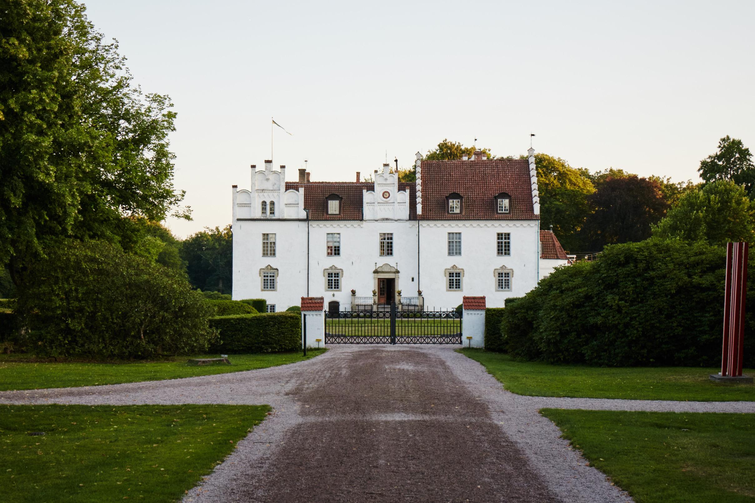 Ein großes weißes Steinhaus hinter einem Eisentor, umgeben von einem grünen Garten mit großen Bäumen.