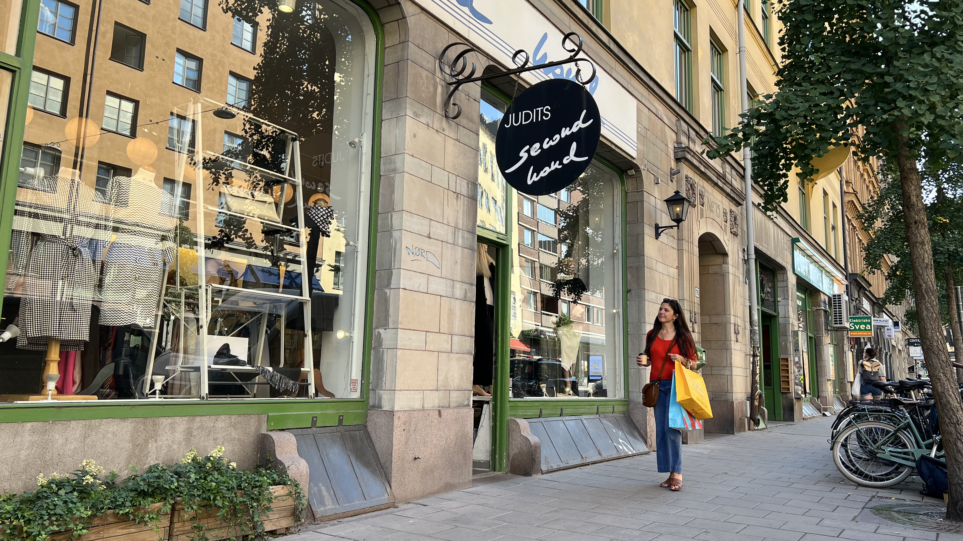 Eine Frau betritt Judits Second Hand in Stockholm an einem sonnigen Tag und trÀgt Einkaufstaschen vor dem Schaufenster.