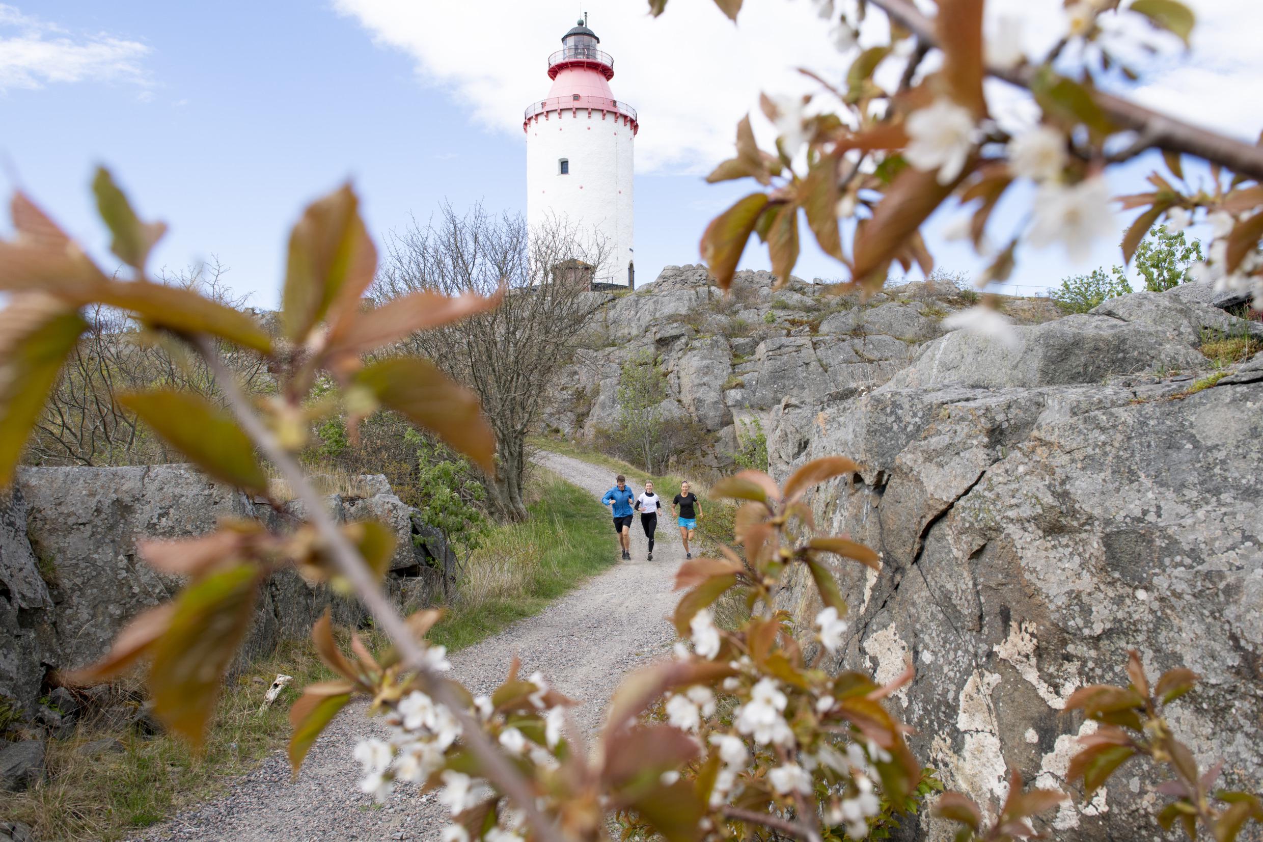 Drei Personen joggen auf einer Schotterstraße. Hinter ihnen steht ein Leuchtturm.