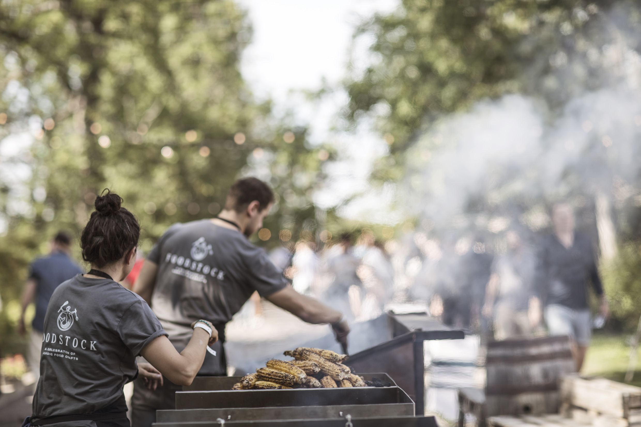 Two people are working outdoors, at a big grill at a festival. A pile of corn cobs lies on the grill.