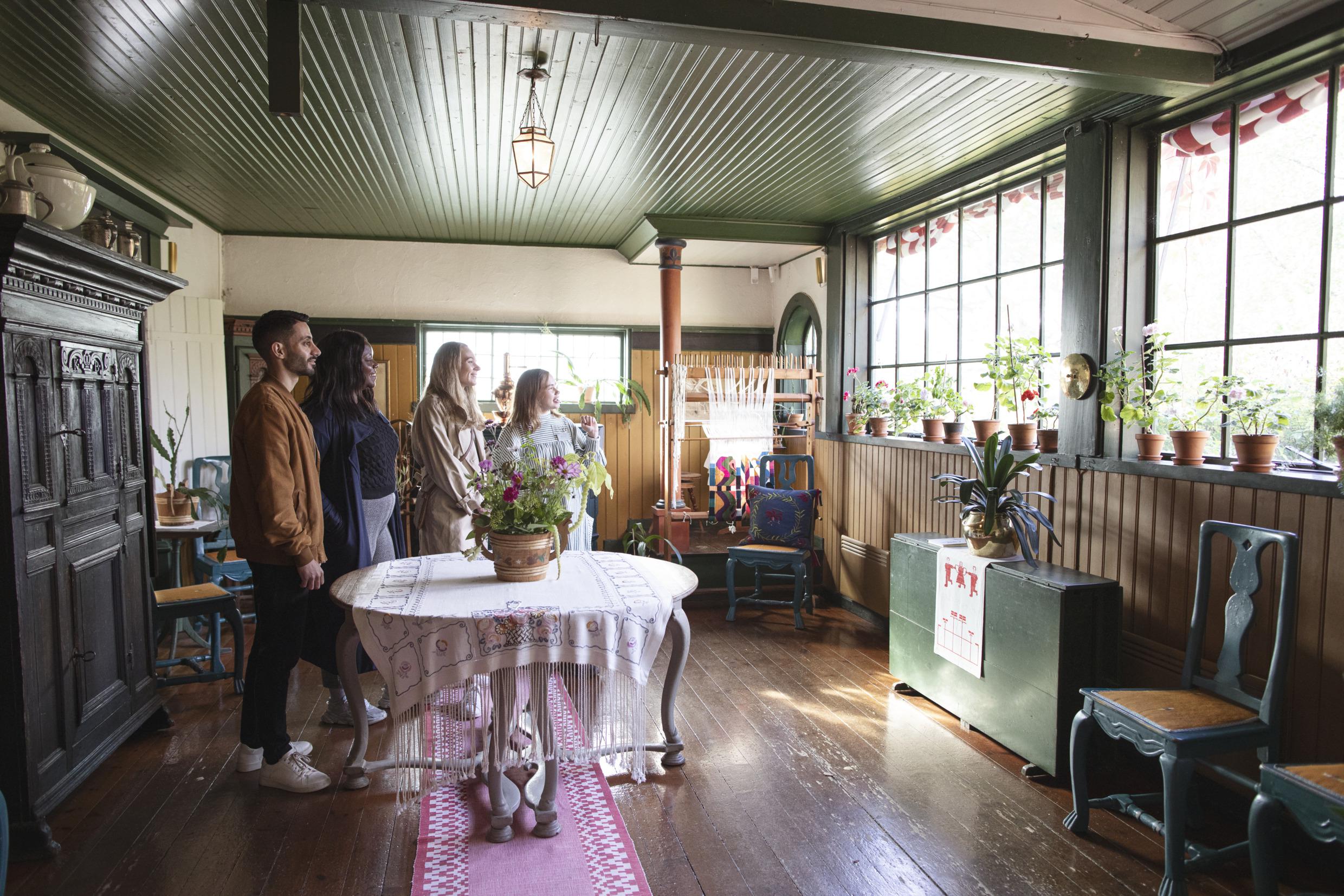 Eine Gruppe von drei Personen hört einem Führer zu, während sie in einem mit Möbeln, Blumen, Tischdecken und bestickten Teppichen dekorierten Raum stehen.