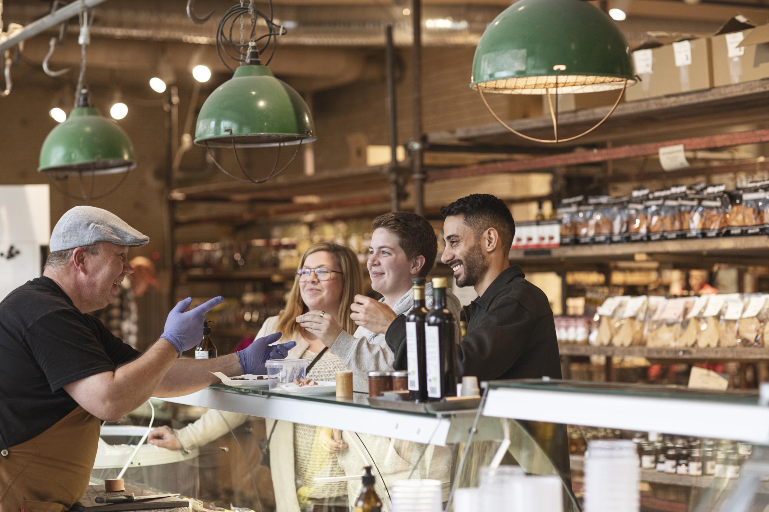 Drei Personen stehen an einer Bedientheke in einer Lebensmittelhalle und sprechen mit einer Verkäuferin.