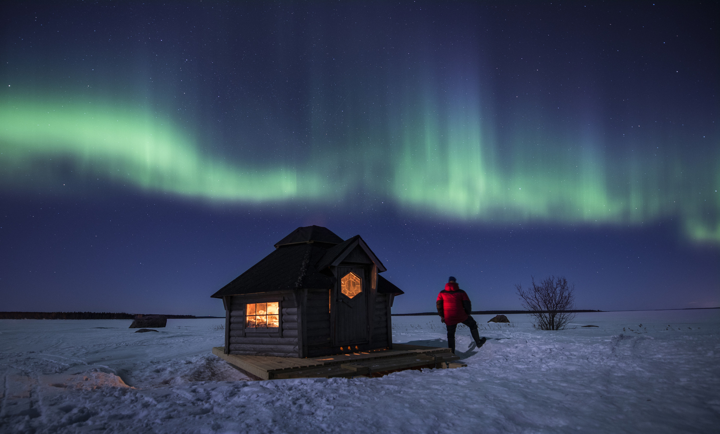 Une personne se tient dans la neige à côté d'une petite maison et regarde les aurores boréales dans le ciel.