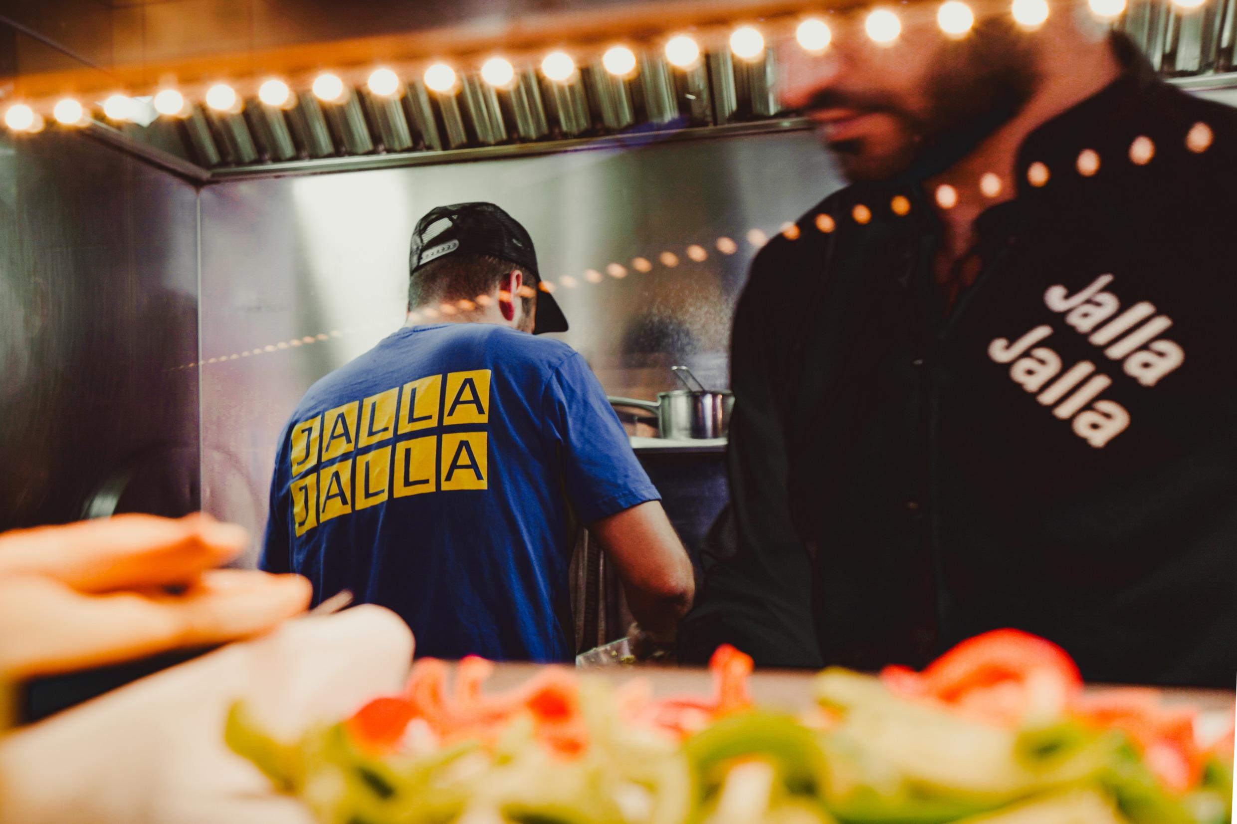 Zwei Männer arbeiten in einer Restaurantküche. Sie tragen T-Shirts mit dem Logo „Jalla Jalla“. Gemüse und Fladenbrot im Vordergrund.