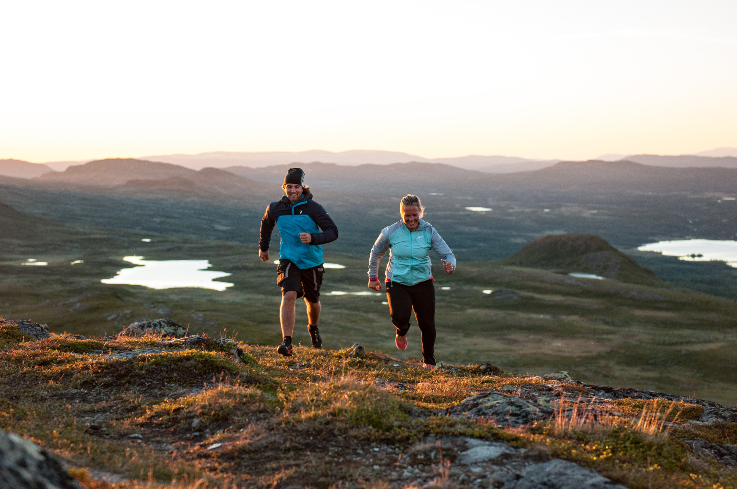 Es sind zwei Läufer zu sehen, die auf einem Bergpfad in Tänndalen in Schweden unterwegs sind. Im Hintergrund sind Seen und sanfte Hügel zu sehen. Die Aufnahme wurde bei Sonnenuntergang gemacht.