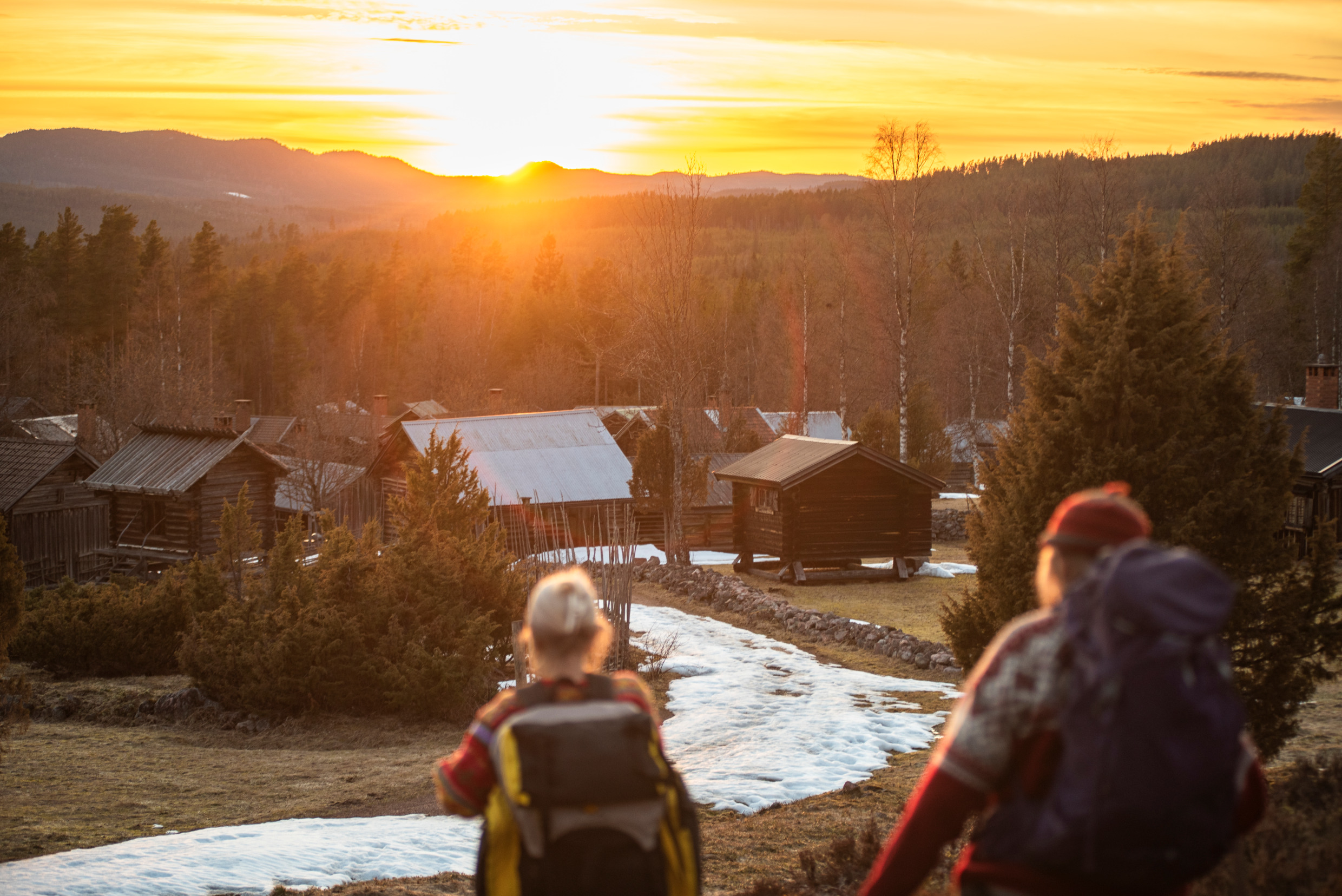 Two persons hikeing while the sun sets over the snow-covered mountains.