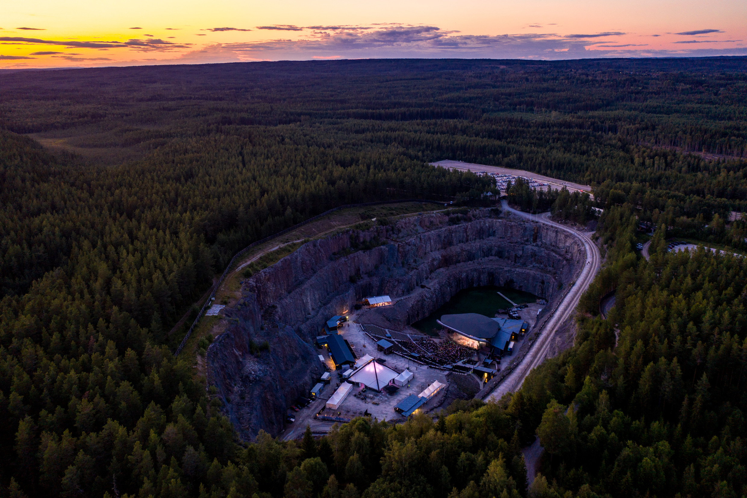 Aerial view of Dalhalla open-air concert venue in a former quarry, surrounded by dense forest and illuminated at dusk in Dalarna, Sweden.