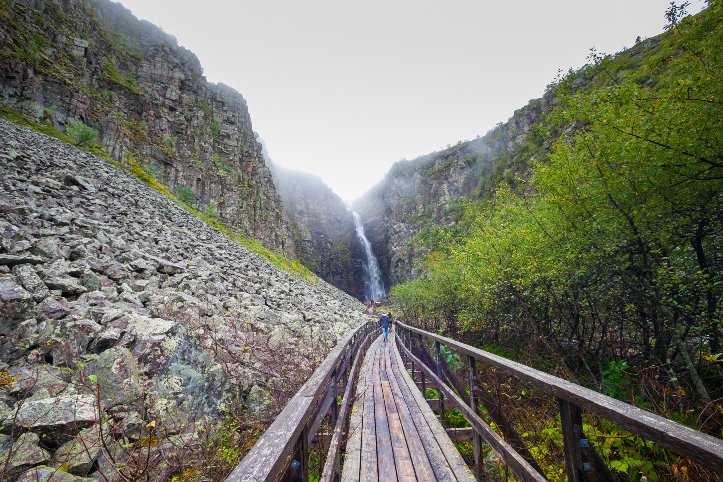 Mensen wandelen op een promenade in het Fulufjällets Nationaal Park, met uitzicht op een waterval.