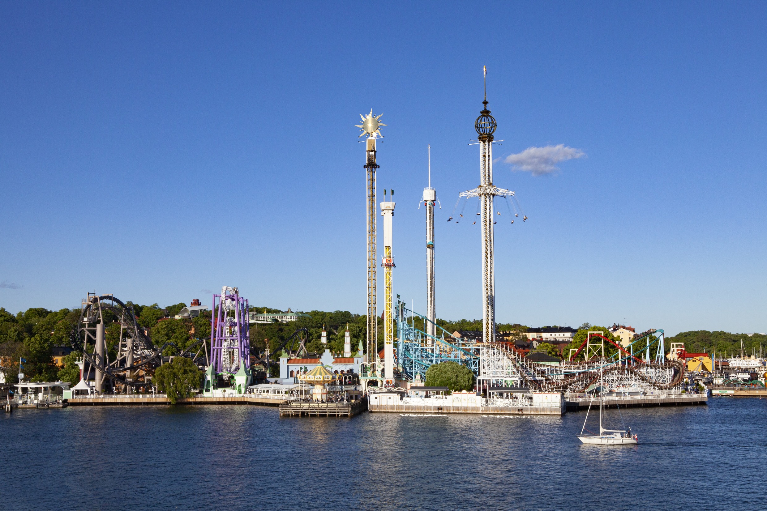 View of Gröna Lund amusement park from the water, showing rollercoasters, tall rides and a sailing boat in the foreground on a sunny day.