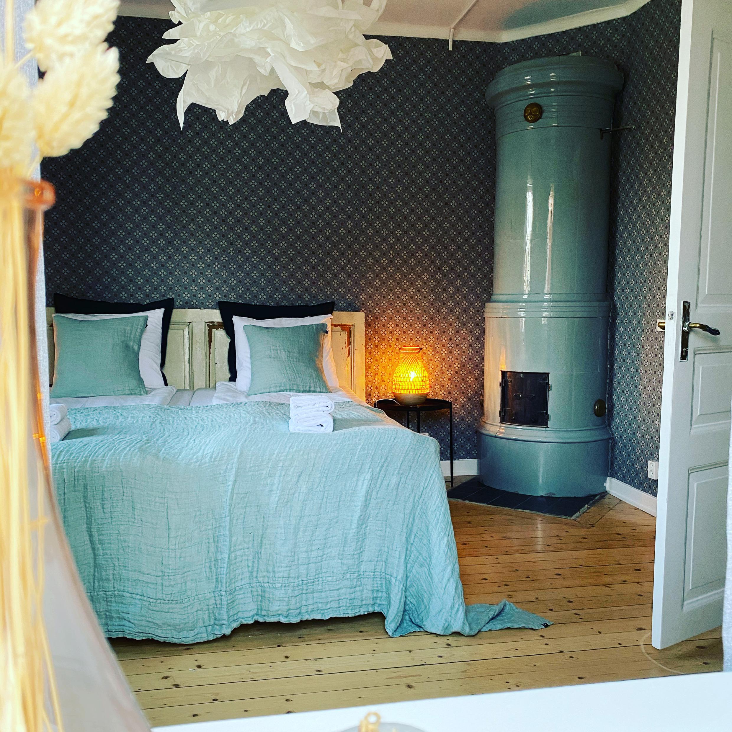 Een hotelslaapkamer met turkoois en wit beddengoed. De muren zijn behangen en in de hoek van de kamer staat een tegelkachel.