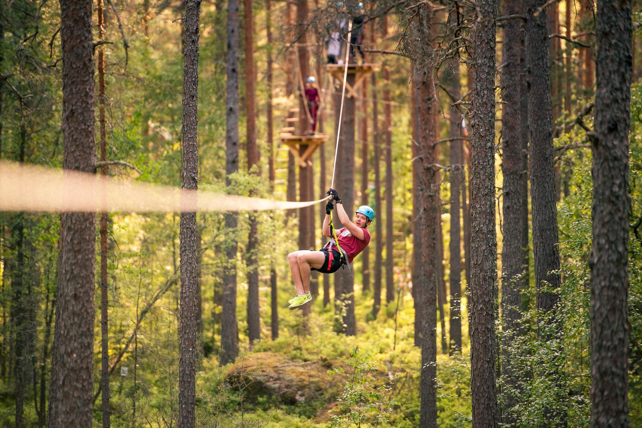Een man is aan het zip-linen in een bos. Aan weerszijden van het tokkelpad staan pijnbomen.