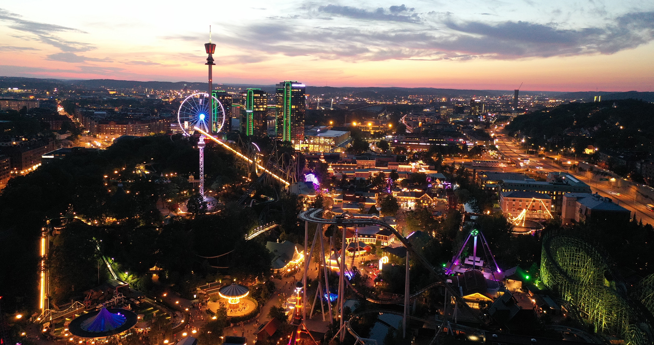 Aerial view of Liseberg amusement park at dusk, with illuminated rides, rollercoasters and city lights in the background.
