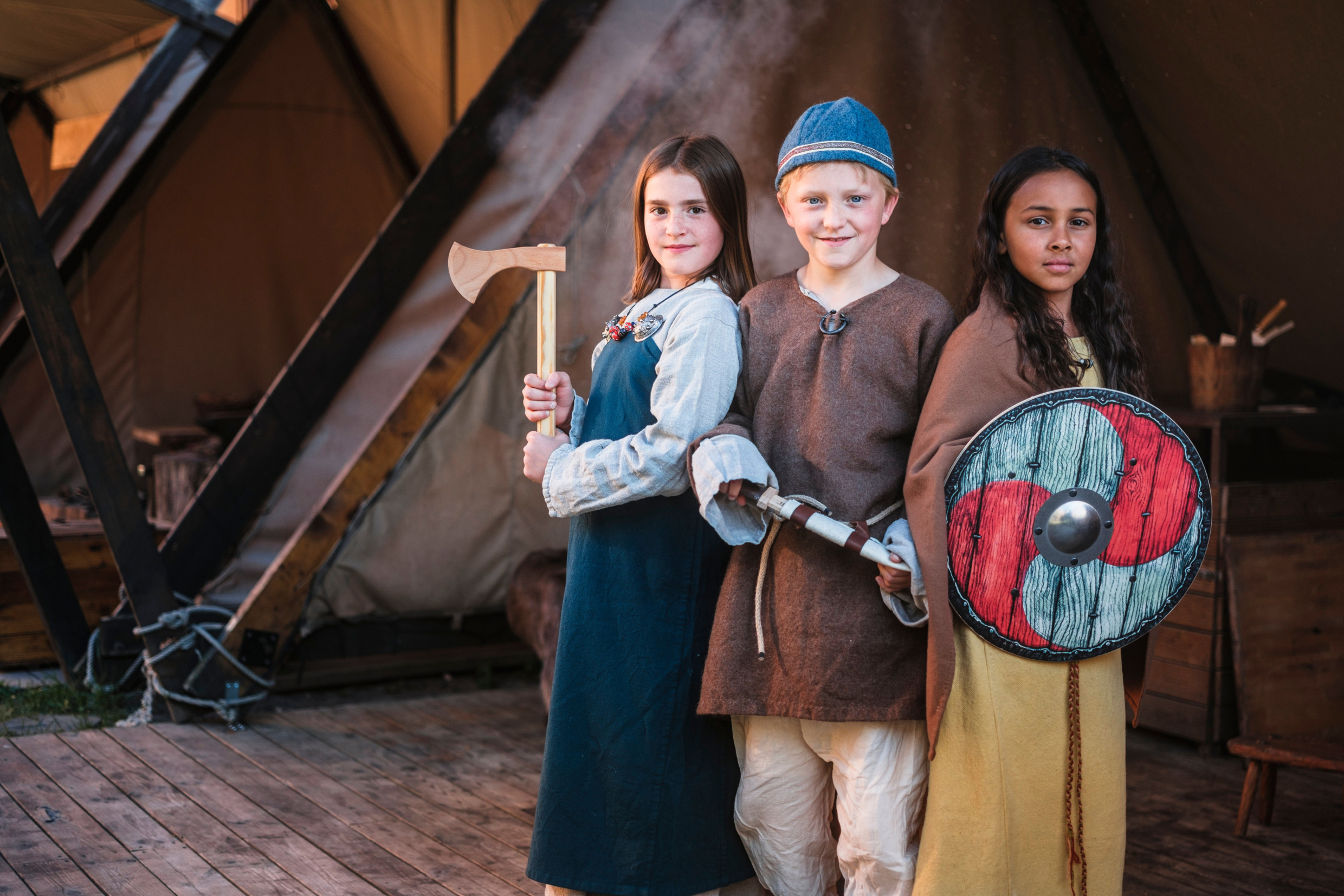 Three children in Viking costumes holding a wooden axe, sword and shield, standing in front of a tent at The Swedish History Museum in Stockholm.