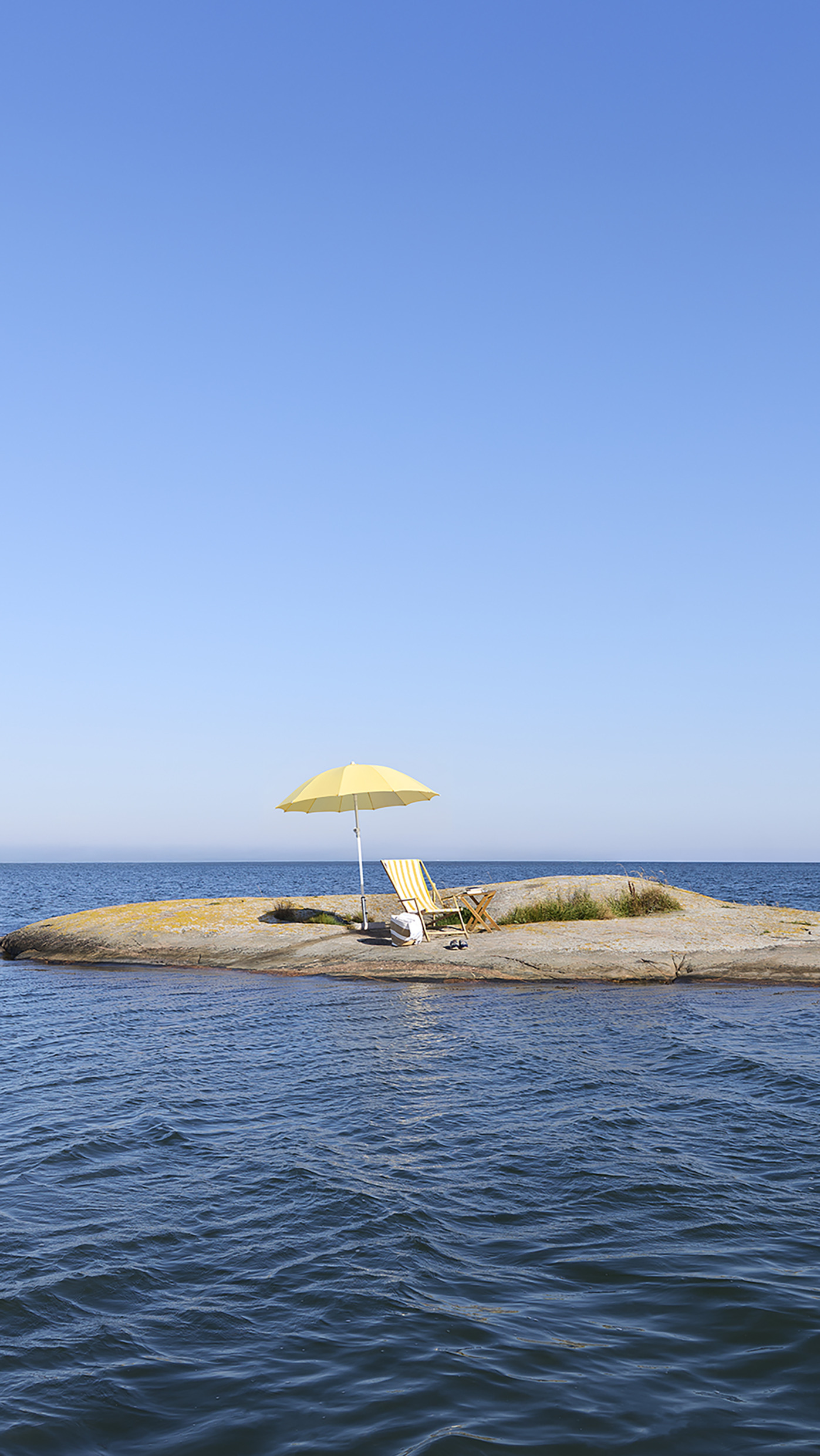A yellow beach umbrella, a sun chair, and a pair of slippers sit on a tiny Swedish islet, surrounded by open sea beneath a clear blue sky.