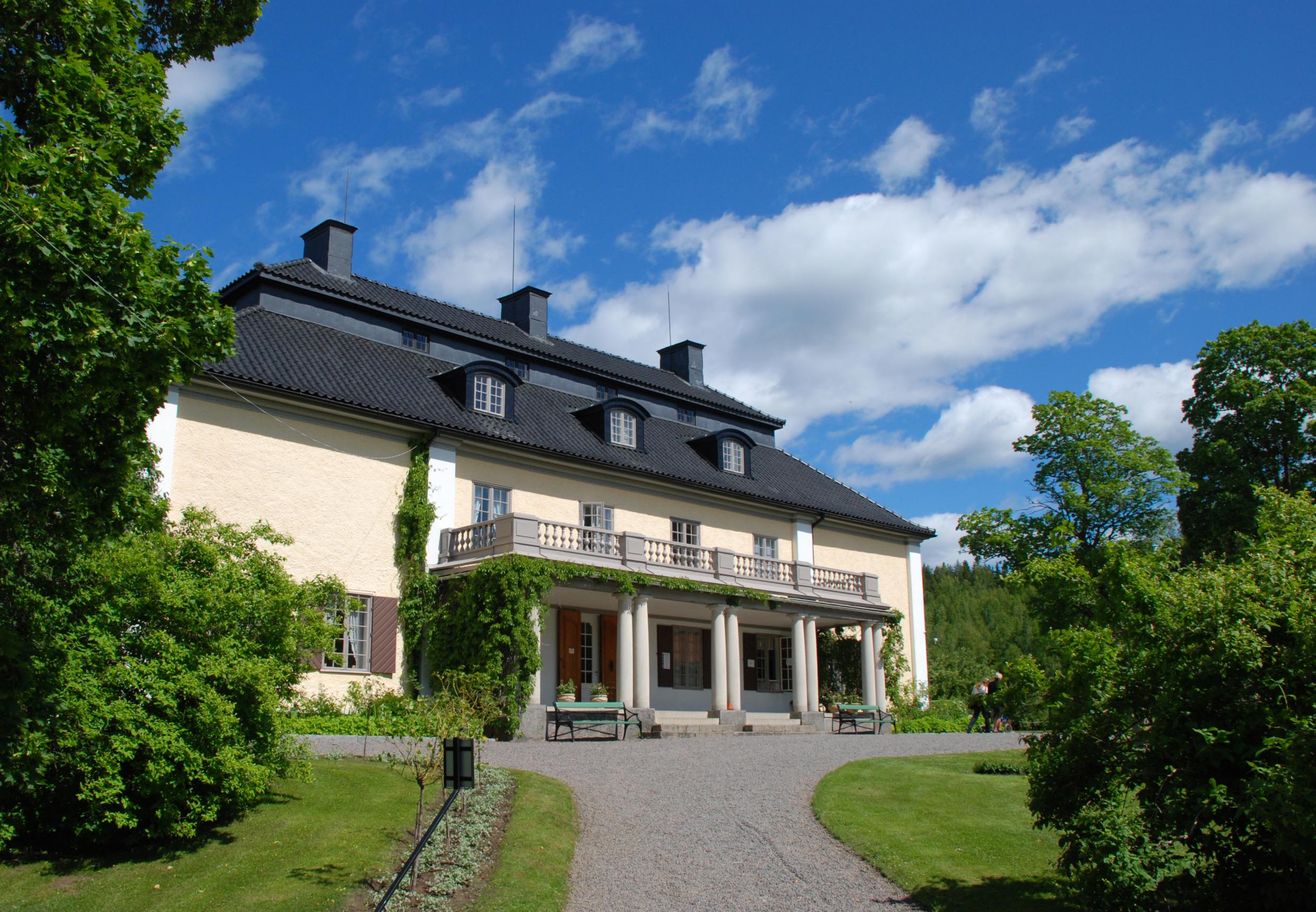 A beautiful light yellow stone house with a garden. In front of the house a green lawn and there are trees and bushes on the sides.