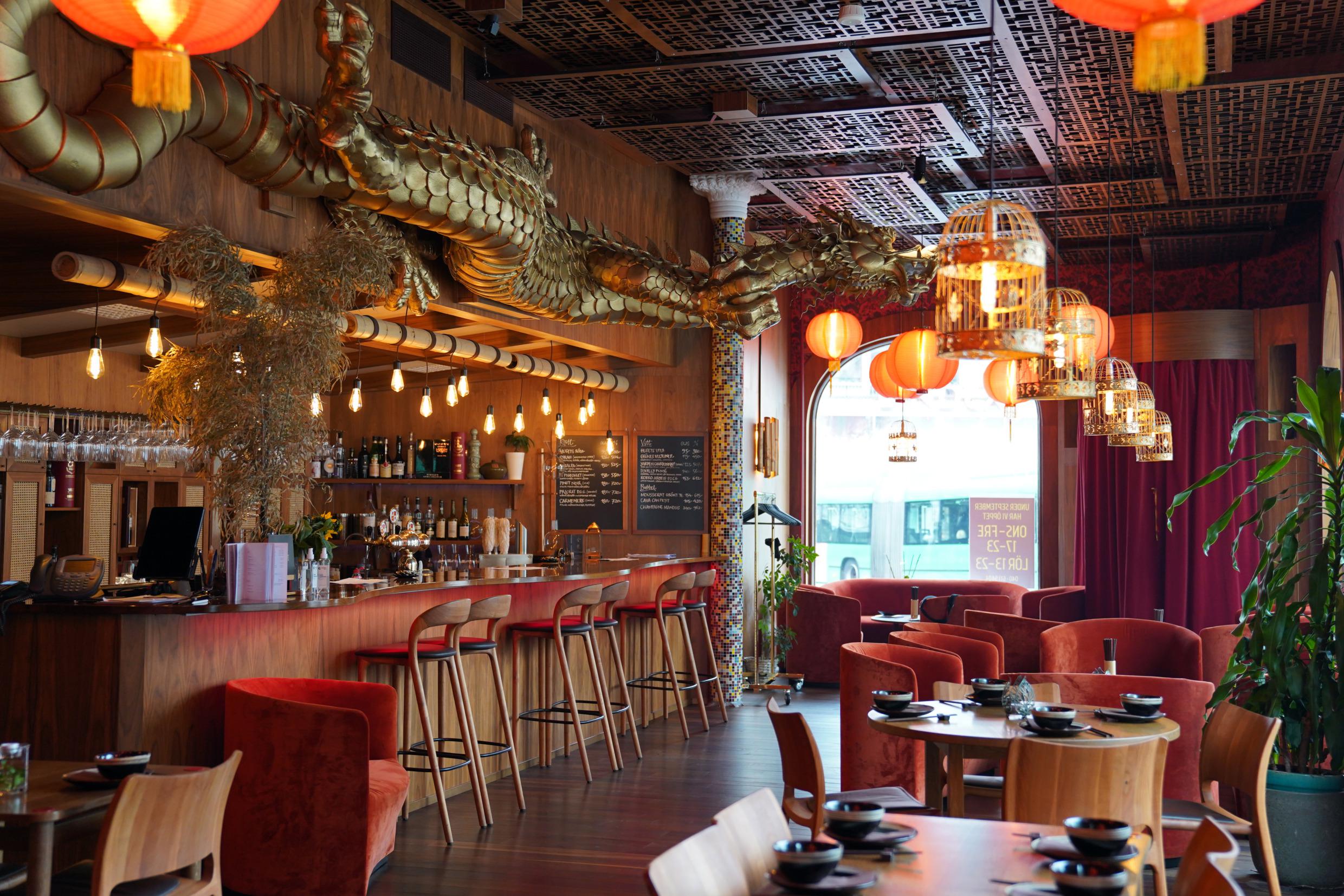 Binnen in een Chinees restaurant. Links een bar met zes barkrukken. Boven de bar hangt een grote gouden draak. Rechts tafels en stoelen. De tafels zijn gedekt met borden, kleine kommen en eetstokjes. Aan het plafond hangen oranje lampen.