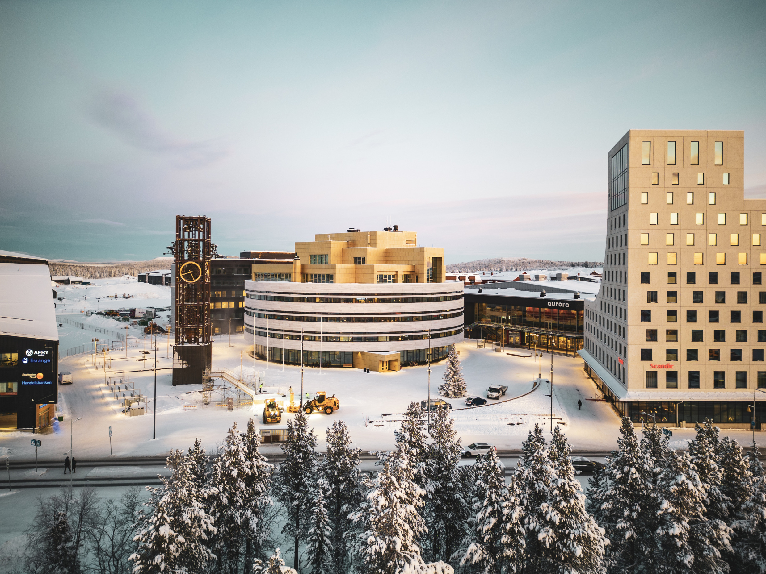 Central Kiruna in winter, with snow-covered trees, the clock tower and modern buildings under a pale sky.