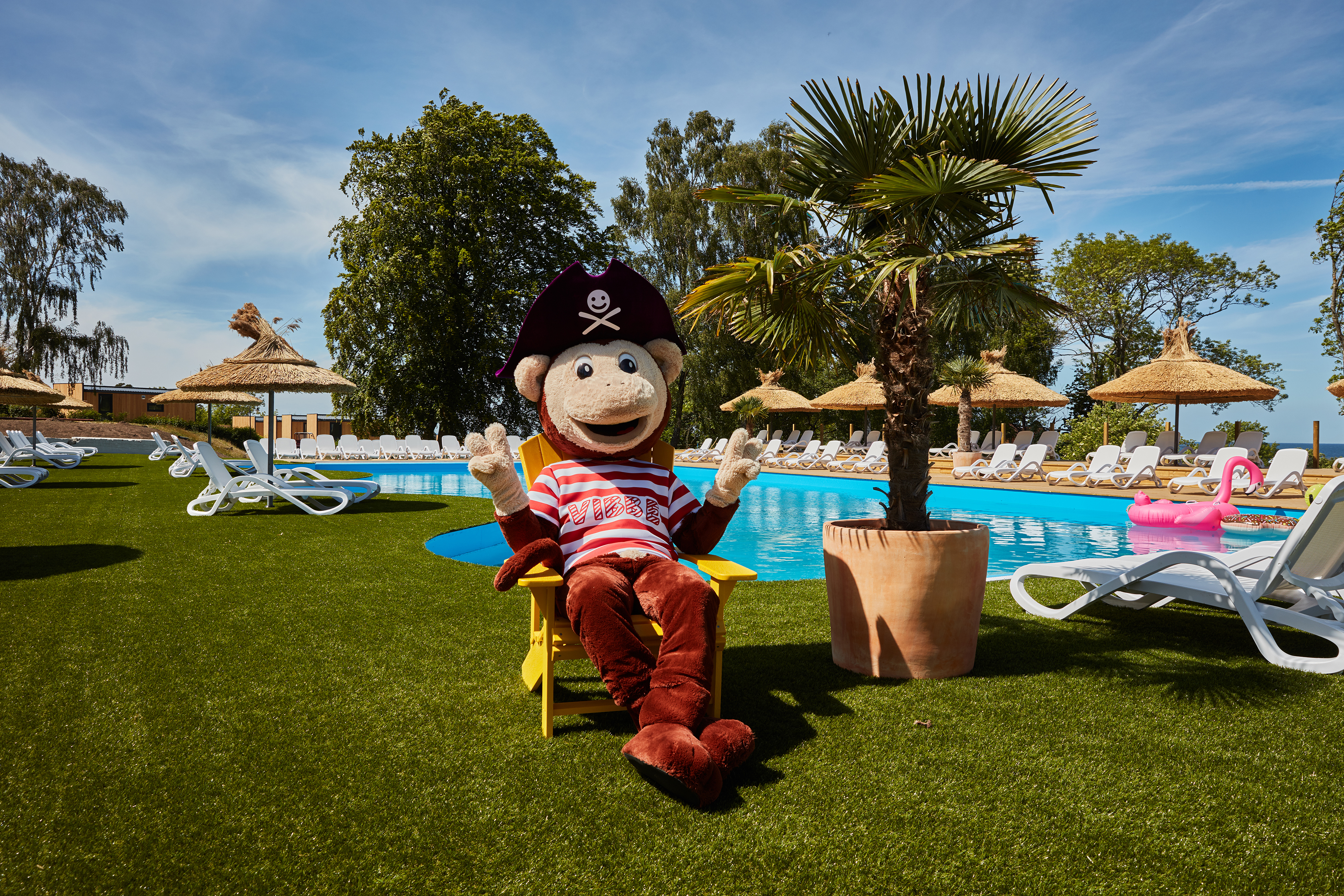Mascot by a pool at Kneippbyn Resort on Gotland with sun loungers and palm trees.