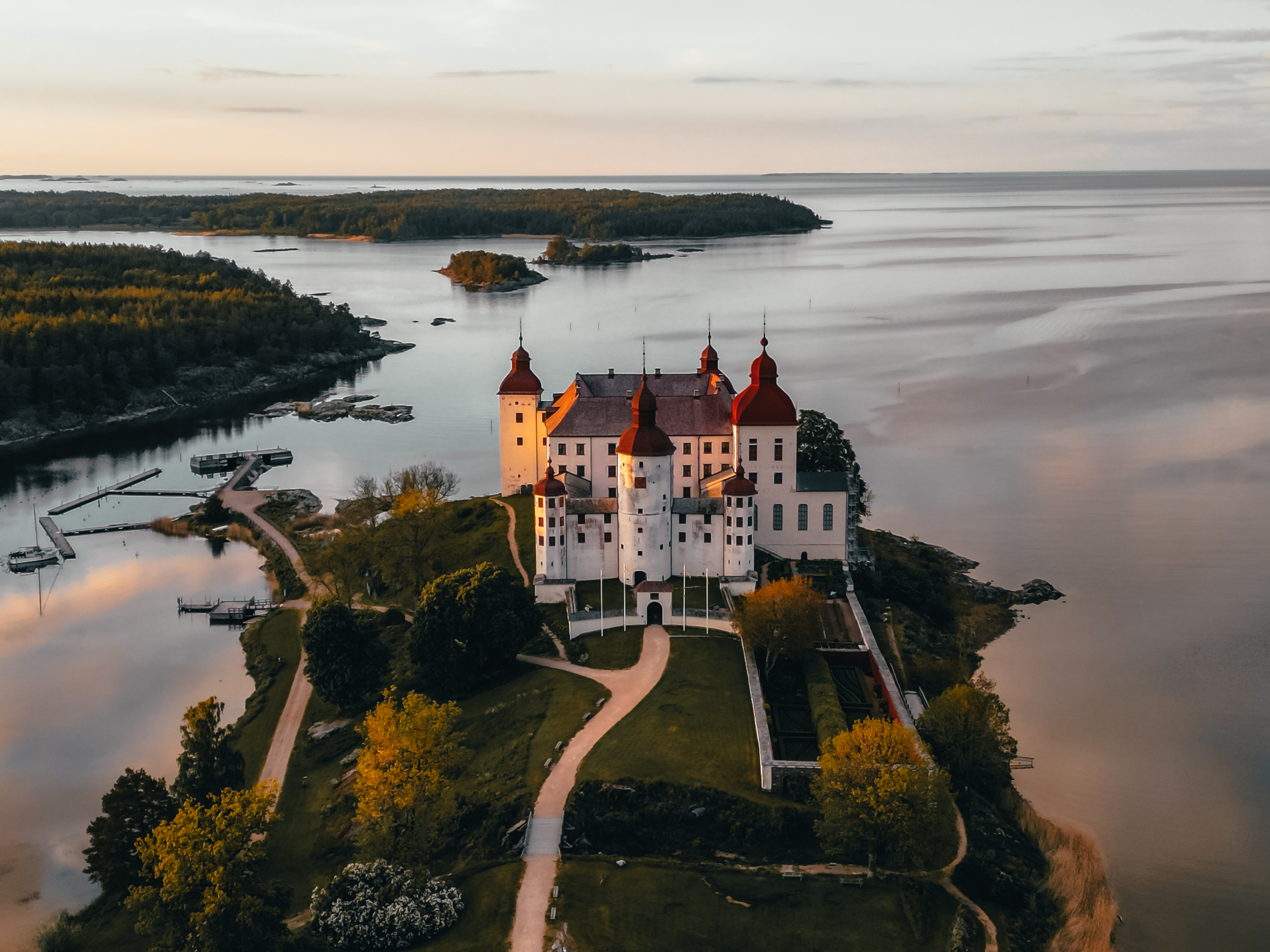 Aerial view of Läckö Castle (Läckö slott) on a peninsula in Lake Vänern, surrounded by calm water, trees and walking paths at sunset.