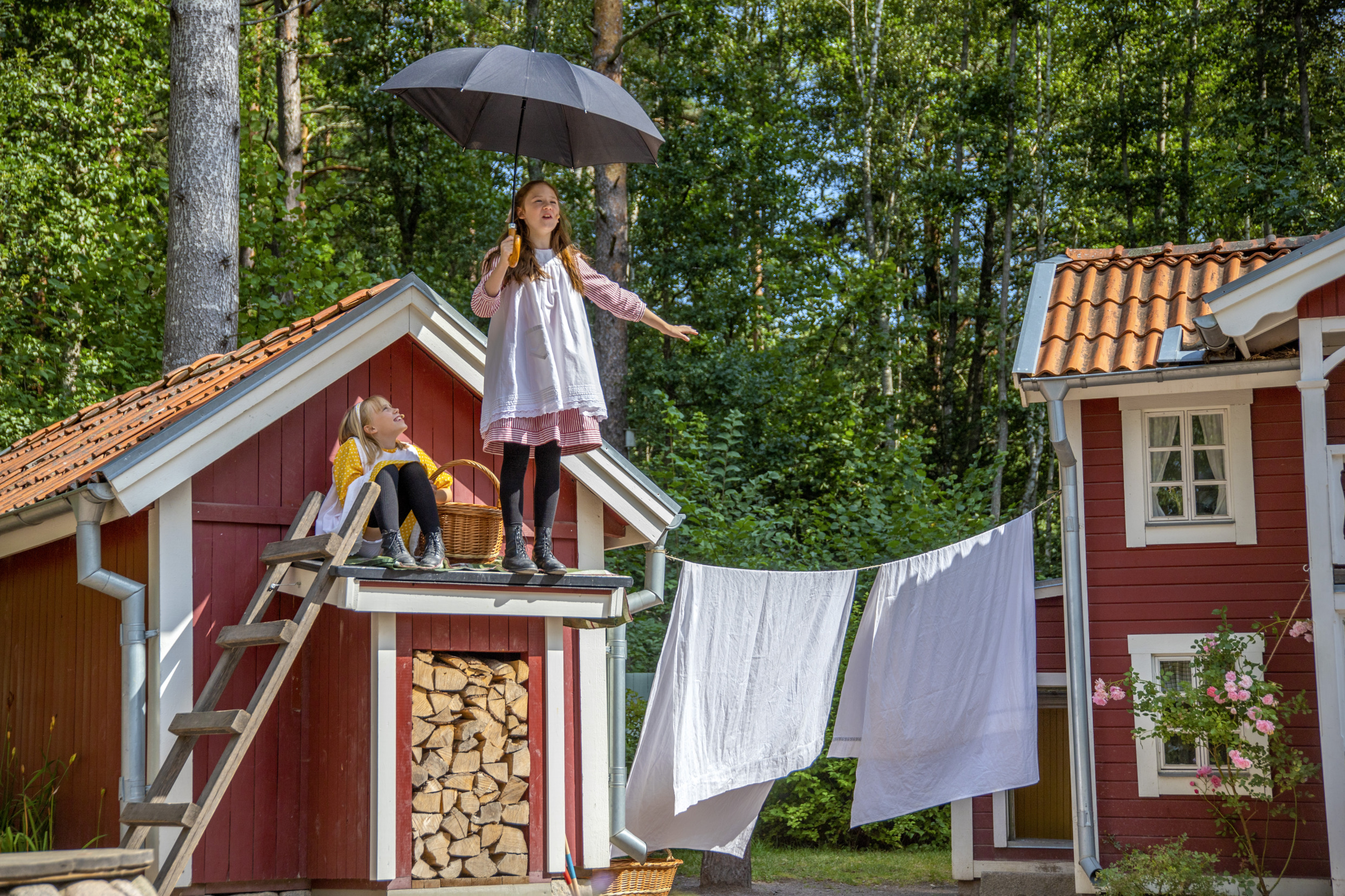 Kinder spielen eine Szene aus „Madicken“ in Astrid Lindgrens Welt in Småland. Ein Mädchen steht mit einem Regenschirm auf einem kleinen roten Holzdach.