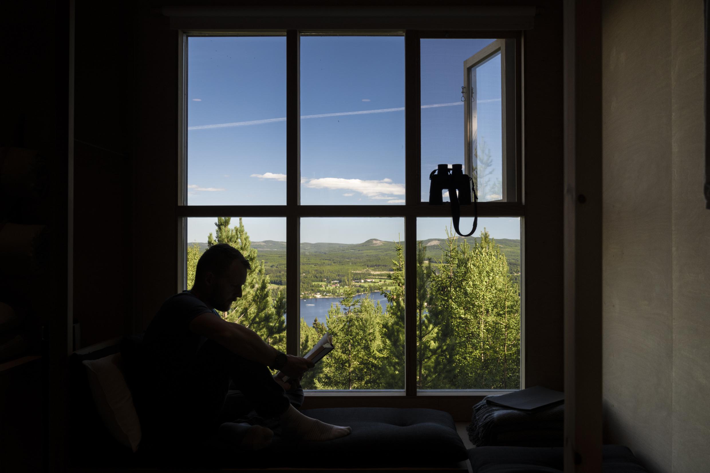 Silhouet van een man die een boek leest bij een groot raam met uitzicht op de blauwe lucht en de bergen in Hälsingland, met een verrekijker bij de hand.