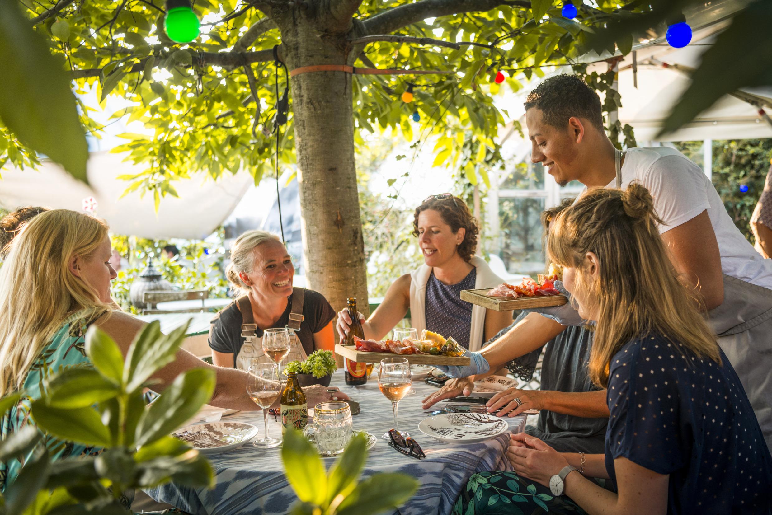 Eine Gruppe von Frauen isst in einem Gartenrestaurant zu Mittag. Ein Mann serviert ihnen zwei Tabletts mit Aufschnitt.
