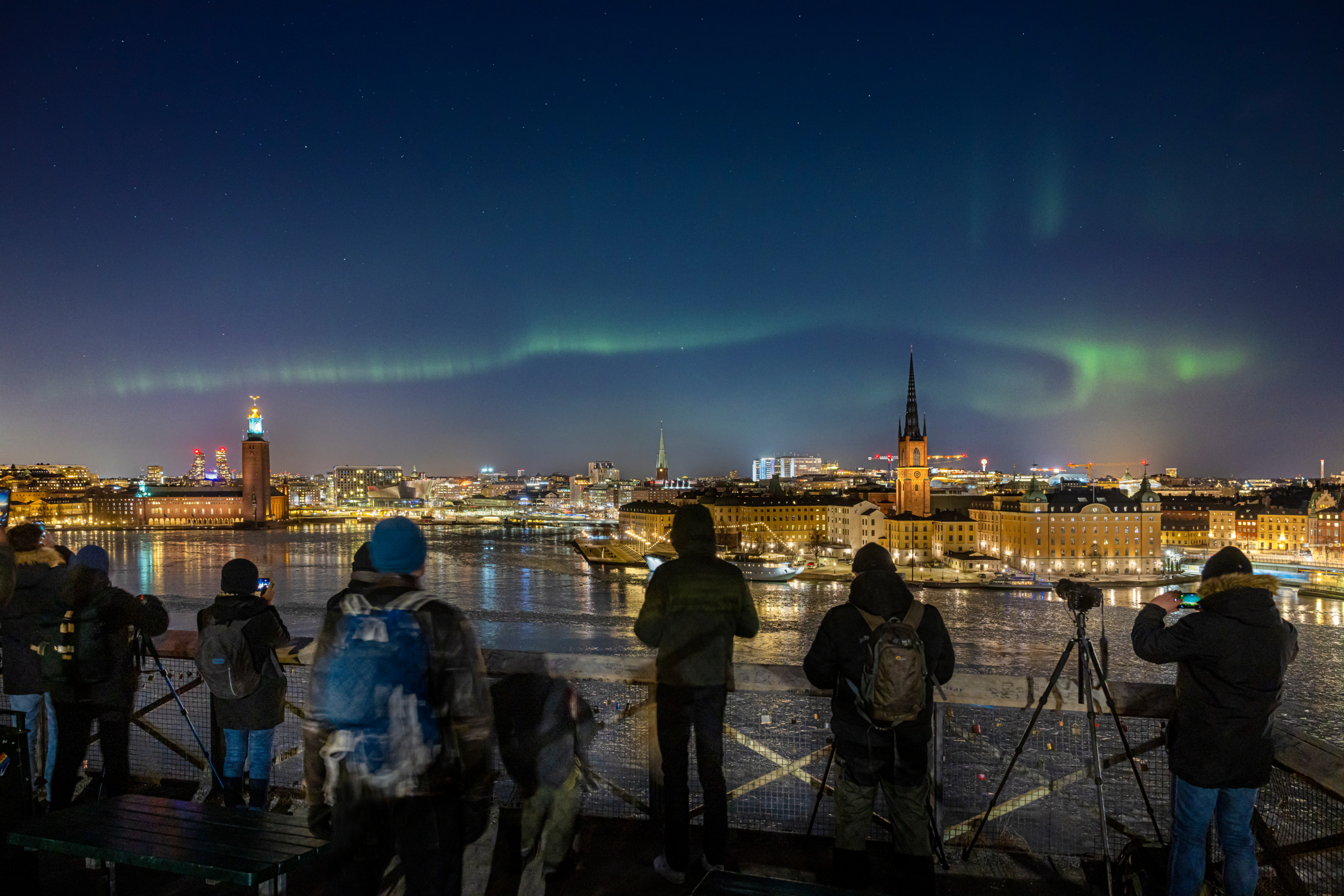 Aurores boréales au-dessus d'un paysage urbain, avec des photographes alignés pour prendre une photo.