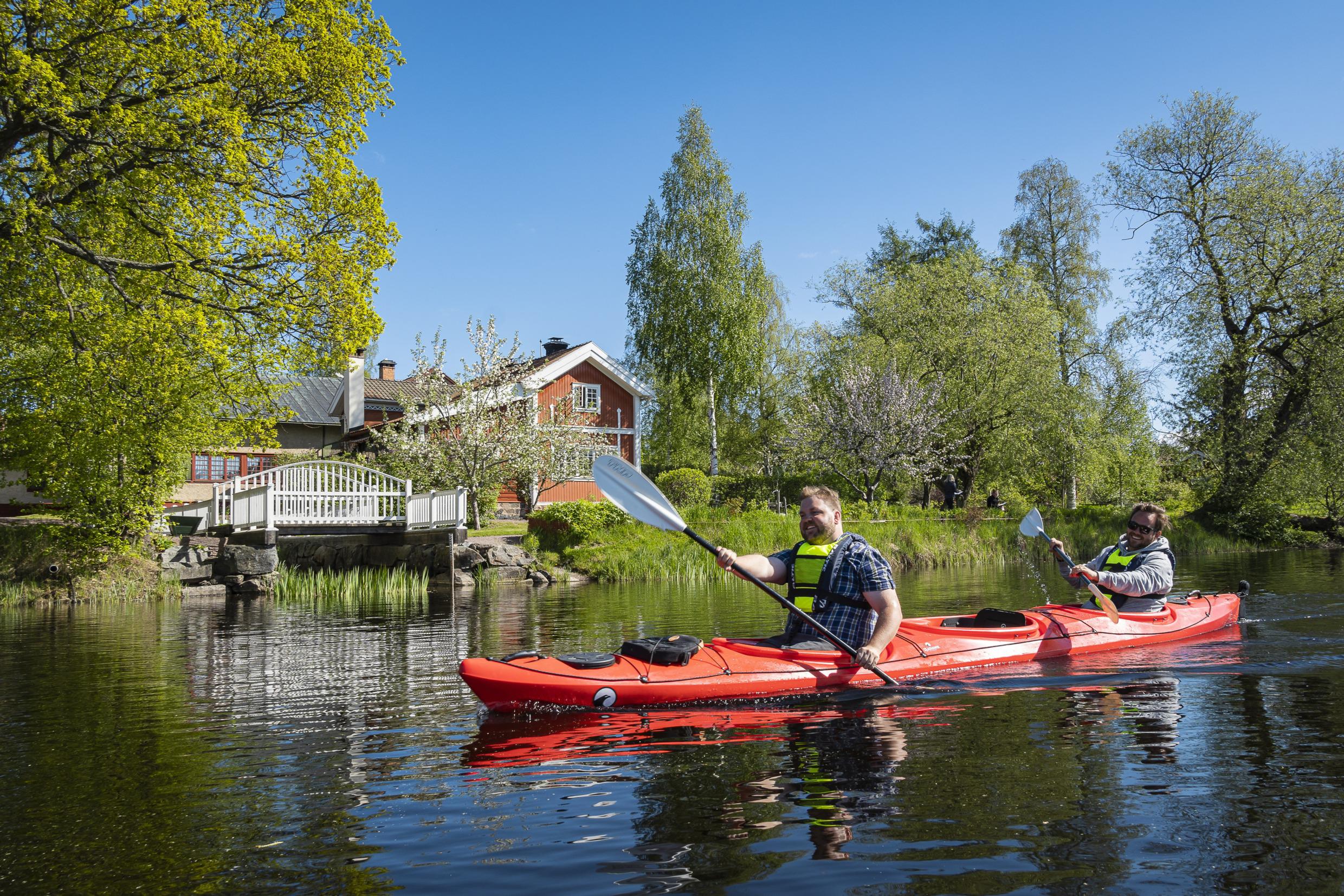 Zwei Männer in einem roten Kajak paddeln neben einem roten Haus mit weißen Zierleisten.