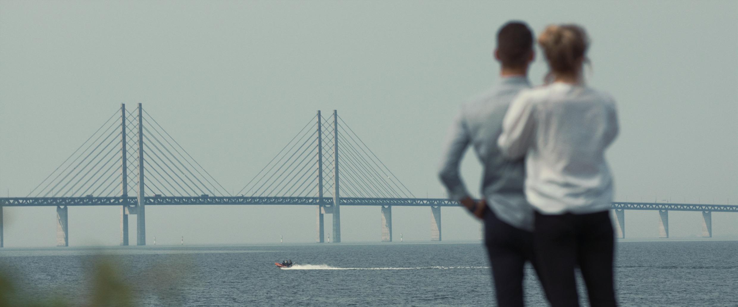 The backs of a man and woman gazing out over the Öresund bridge and a speed boat in the water.