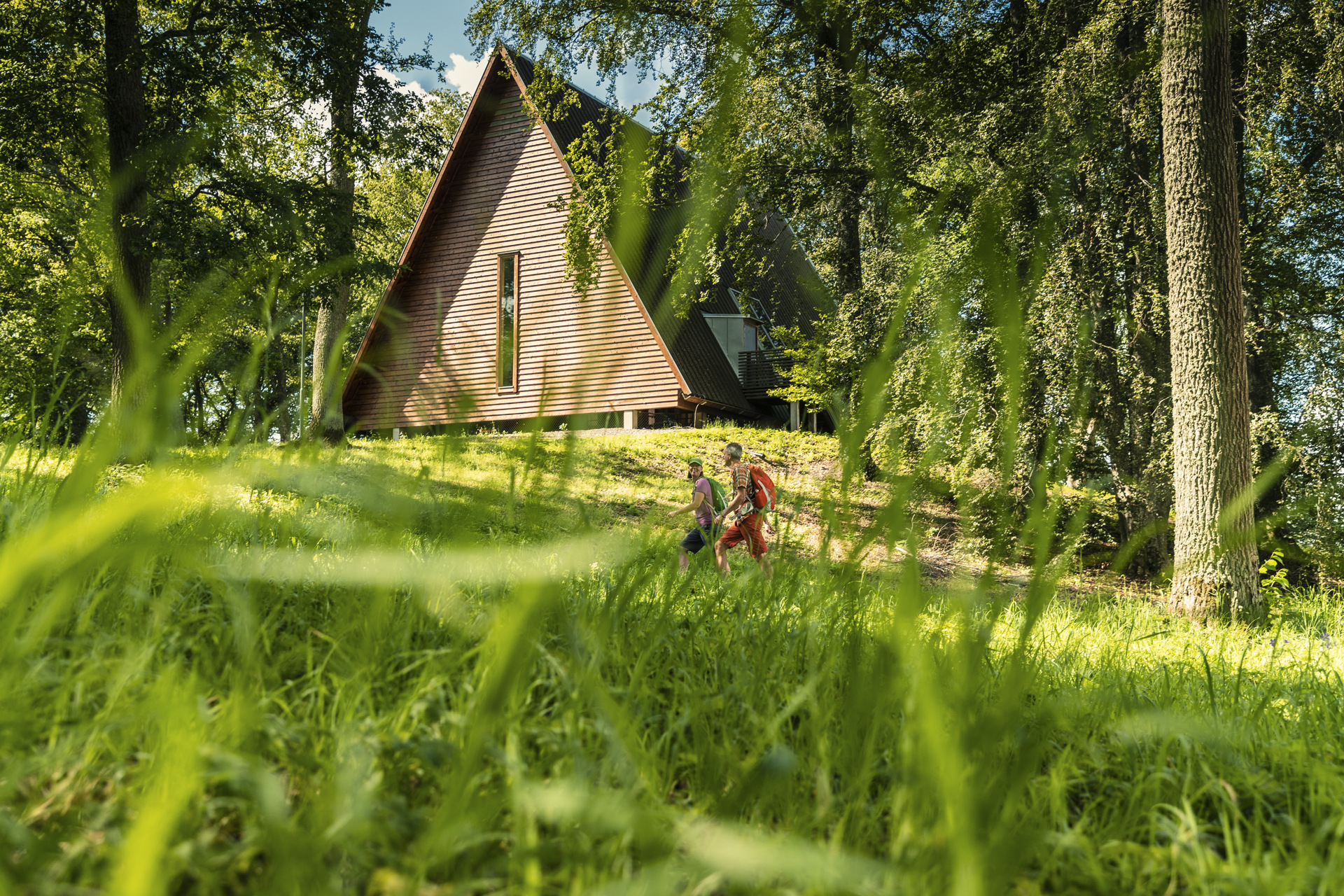 Two hikers walking through a leafy green setting past the wooden Kata Farm building in Varnhem.