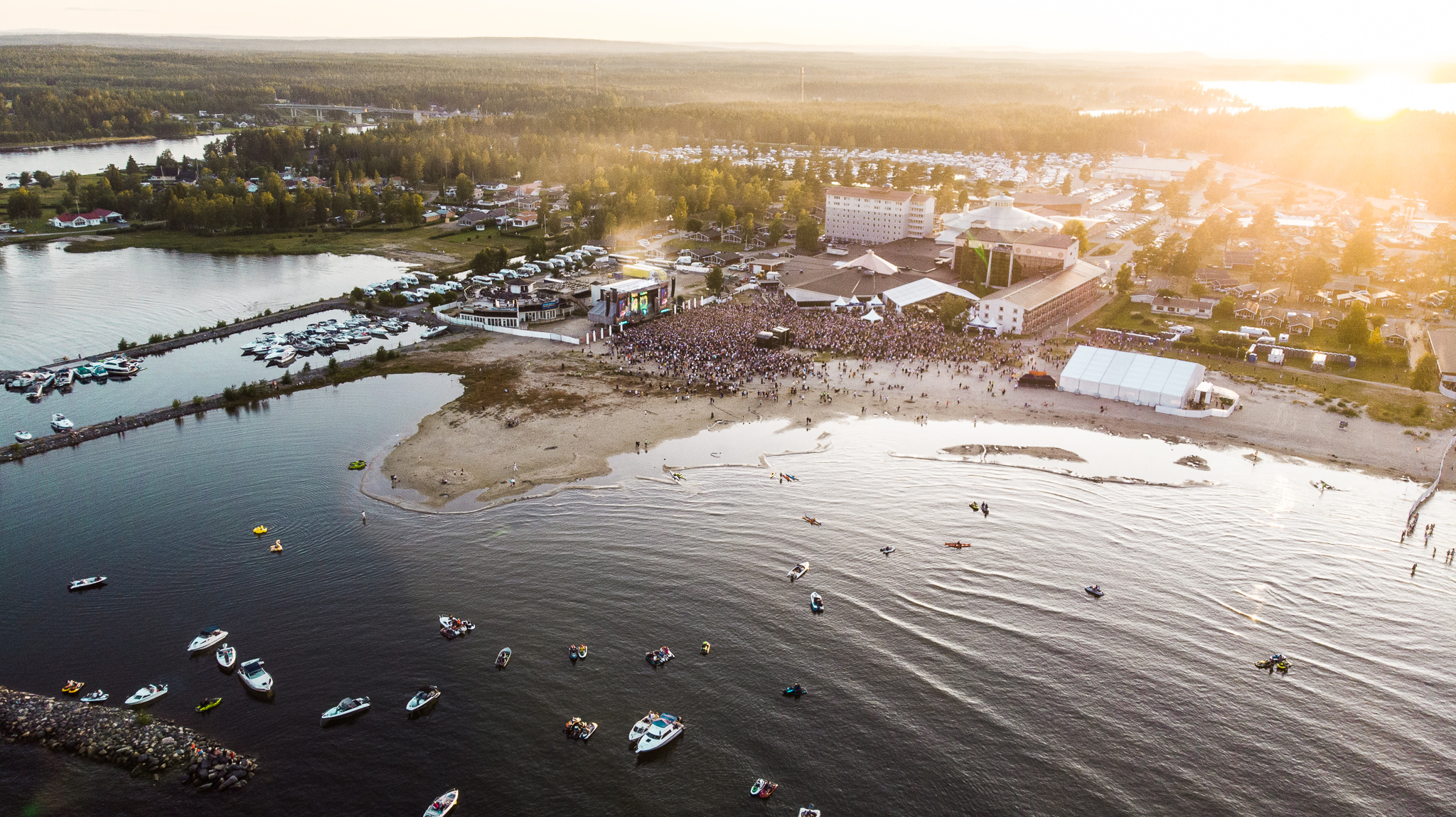 Aerial view of Pite Havsbad with a beach, boats in the water and a large crowd gathered by a stage at sunset.