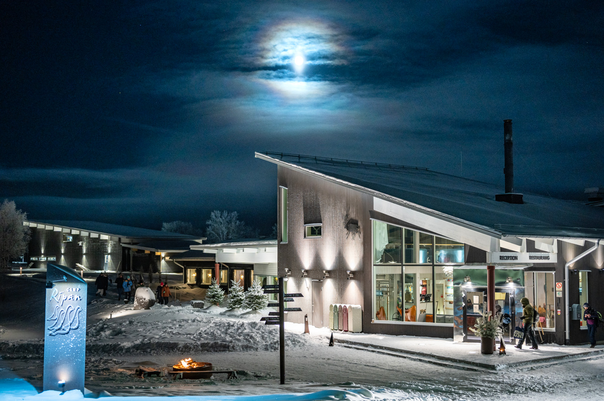 L'entrée enneigée du Camp Ripan à Kiruna, de nuit, avec des bâtiments illuminés, des personnes qui arrivent et un paysage hivernal.