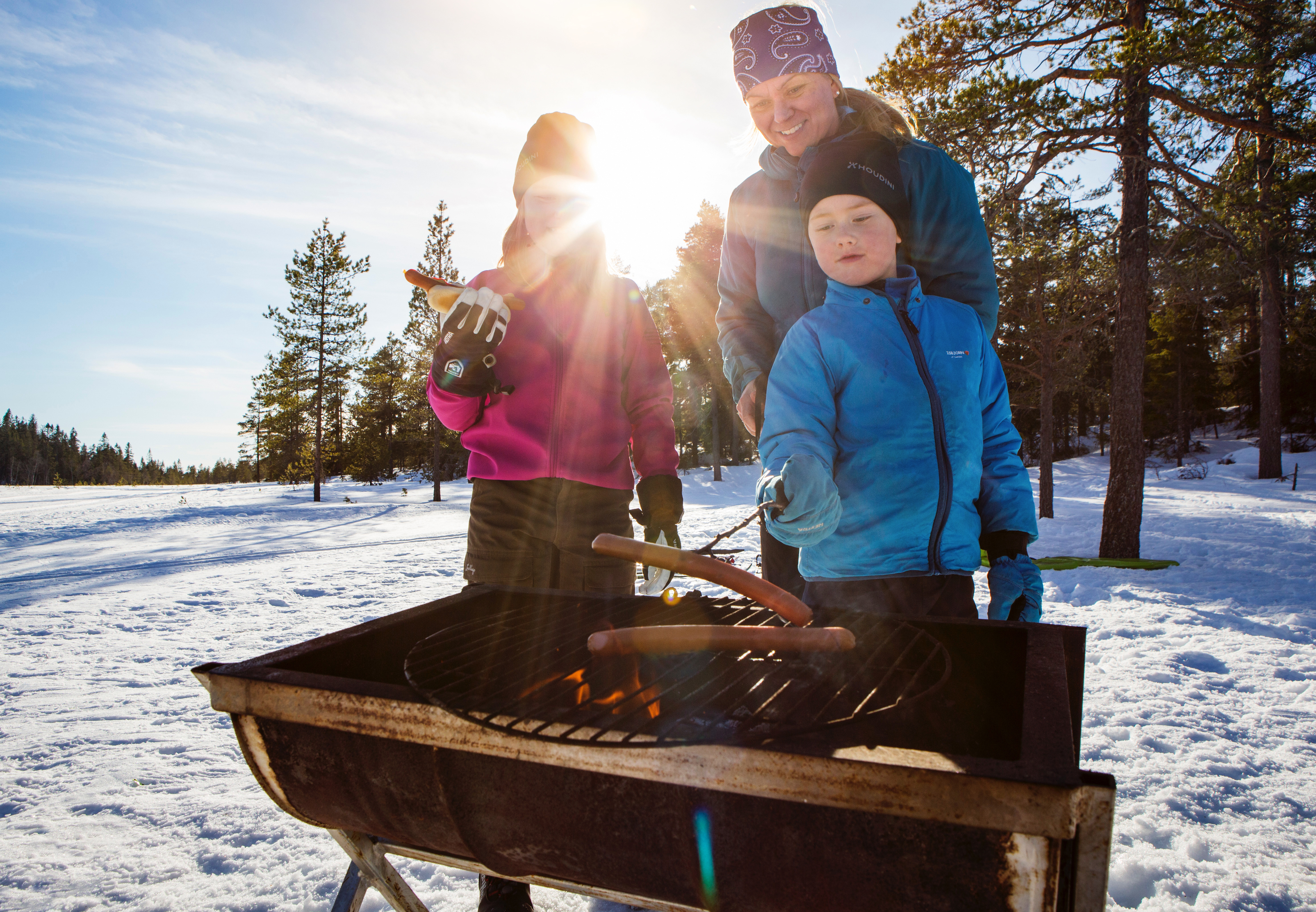 Een familie van 3 personen roostert worstjes boven een barbecue in een winters landschap.