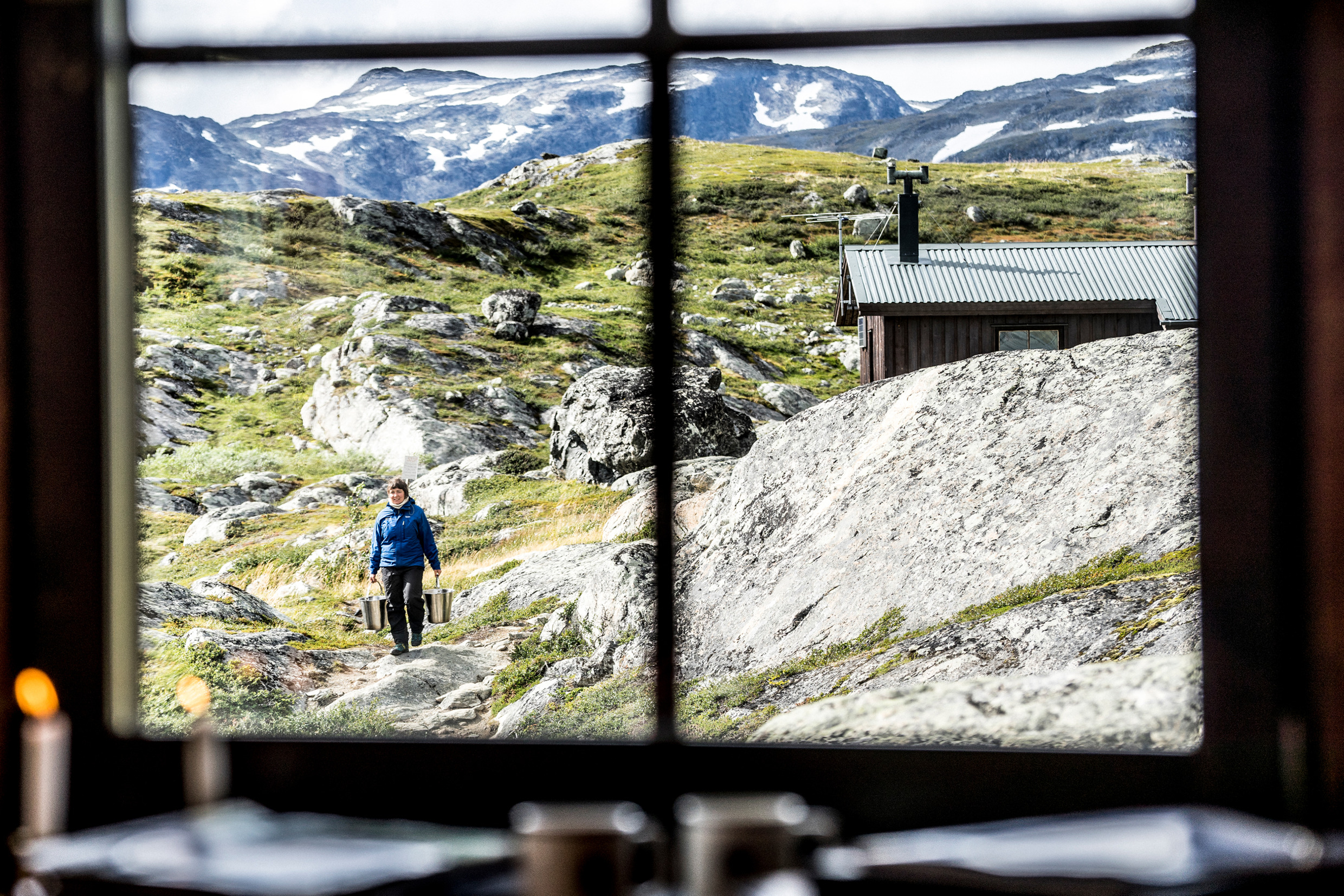 Eine Frau, die durch ein Fenster zu sehen ist, trägt zwei Eimer. Sie geht von einer Holzhütte aus, die Berge liegen hinter ihr