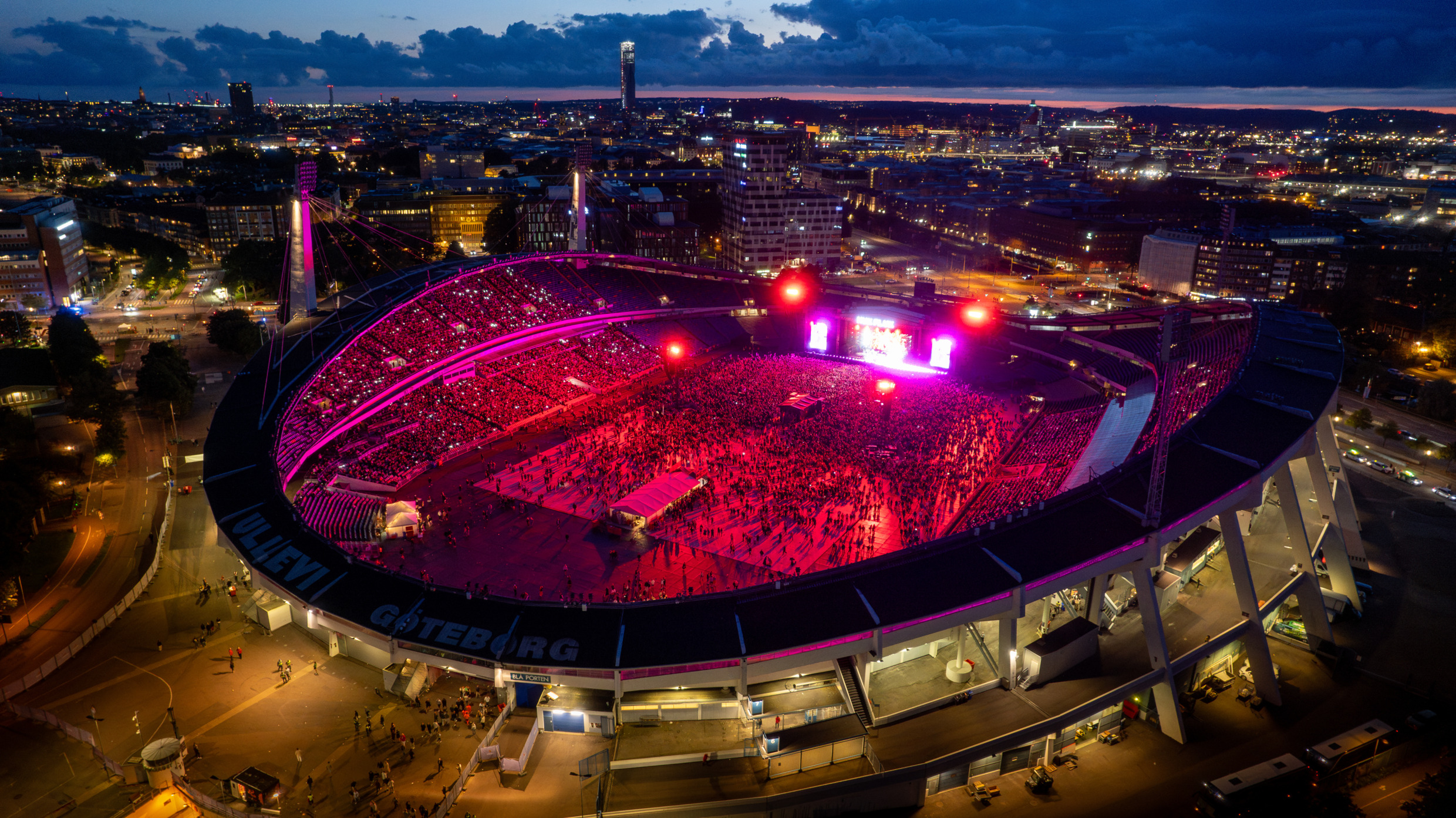 Diese Luftaufnahme zeigt das Ullevi-Stadion in Göteborg während eines Nachtkonzerts mit Bühnenbeleuchtung, vielen Leuten und der Skyline der Stadt im Hintergrund.