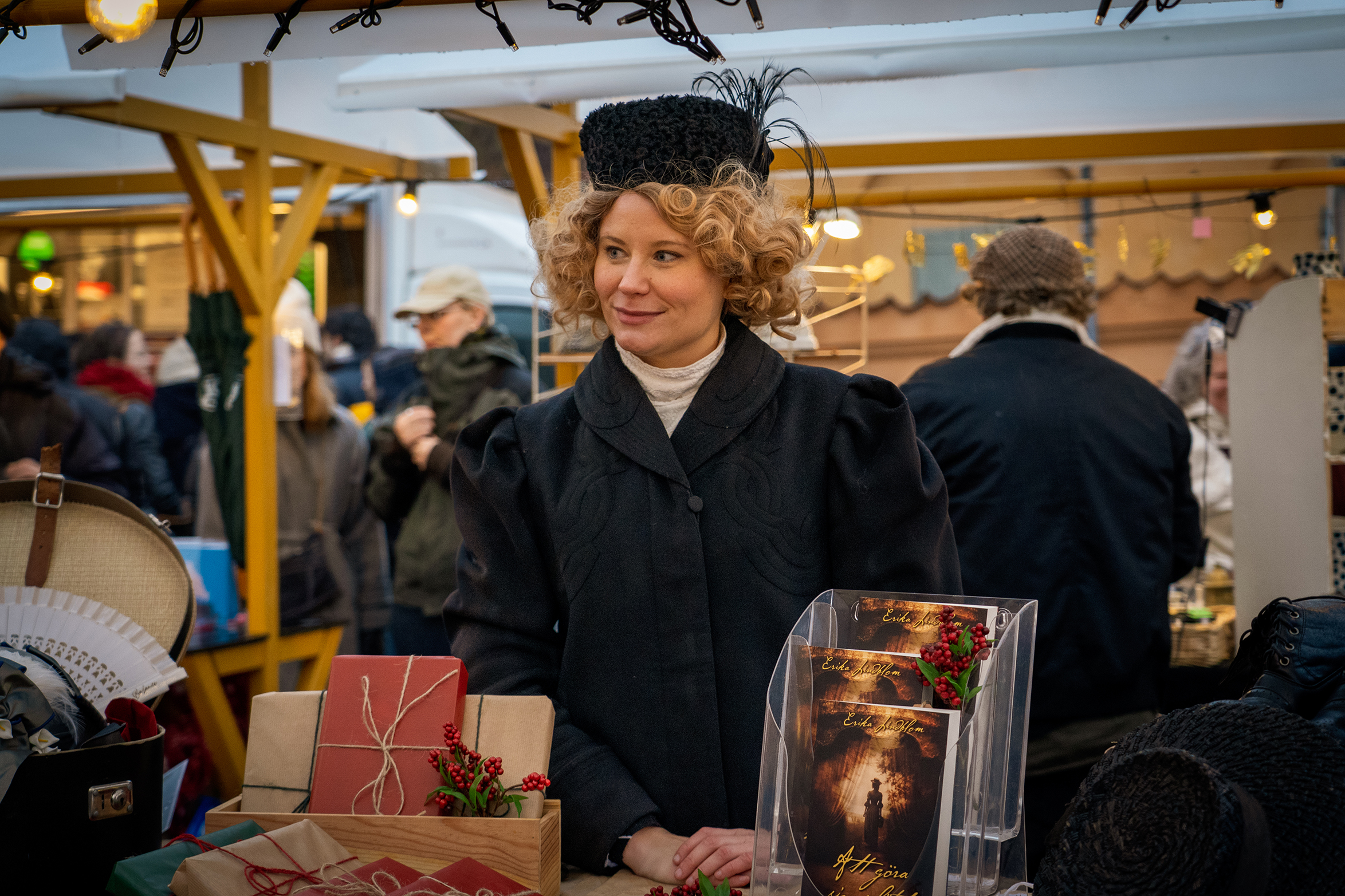 A woman in historical dress stands behind a stall displaying wrapped gifts, books and handmade accessories at Uppsala Christmas Market.