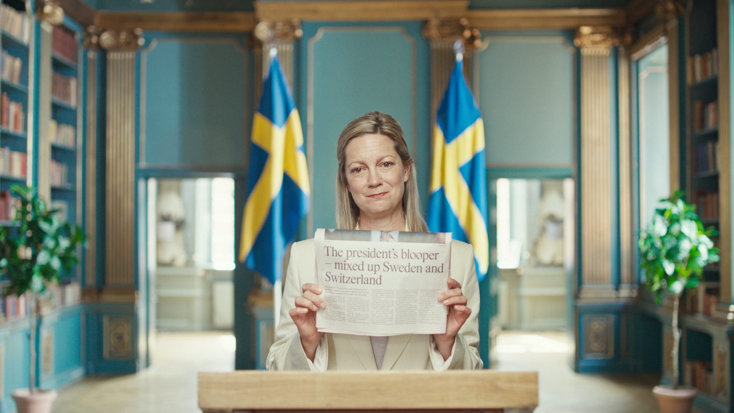 A woman in a white blazer is standing behind a podium, holding a newspaper that reads "President's blooper - mixed up Sweden and Switzerland". Behind her are two Swedish flags.