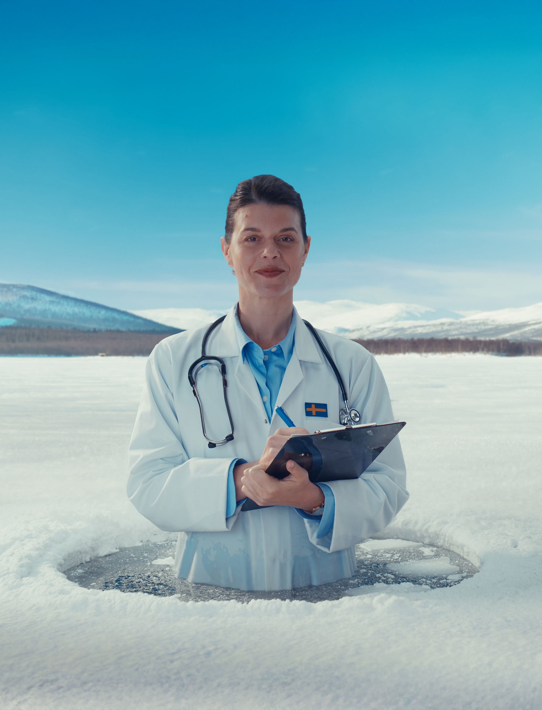 A female doctor stands in a hole in the ice in northern Sweden, surrounded by snow, with mountains rising in the background.