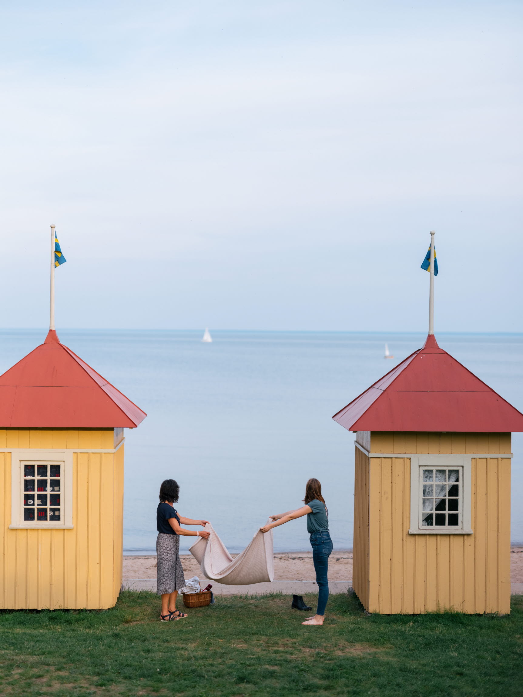 Zwei Frauen breiten eine Picknickdecke vor einer Strandhütte in der Villa Eira in Hjo mit Blick aufs Meer aus.