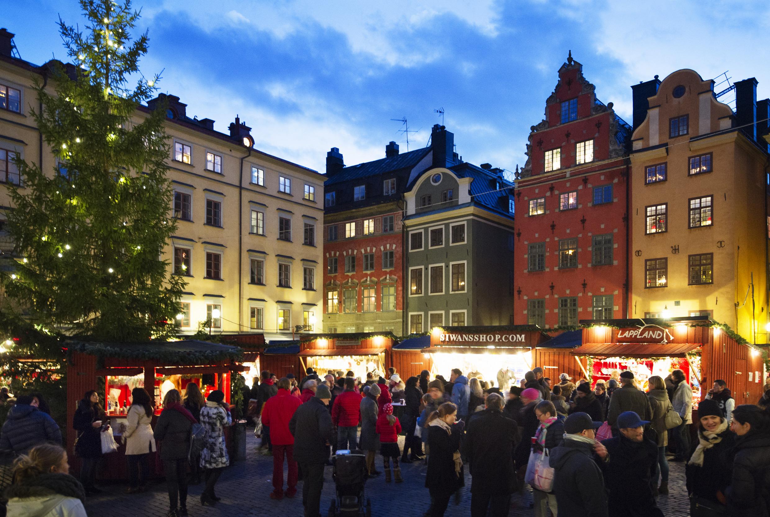 Crowds of people are visiting the stalls of the old-fashioned Christmas market in Old Town Stockholm, next to a lit Christmas tree.