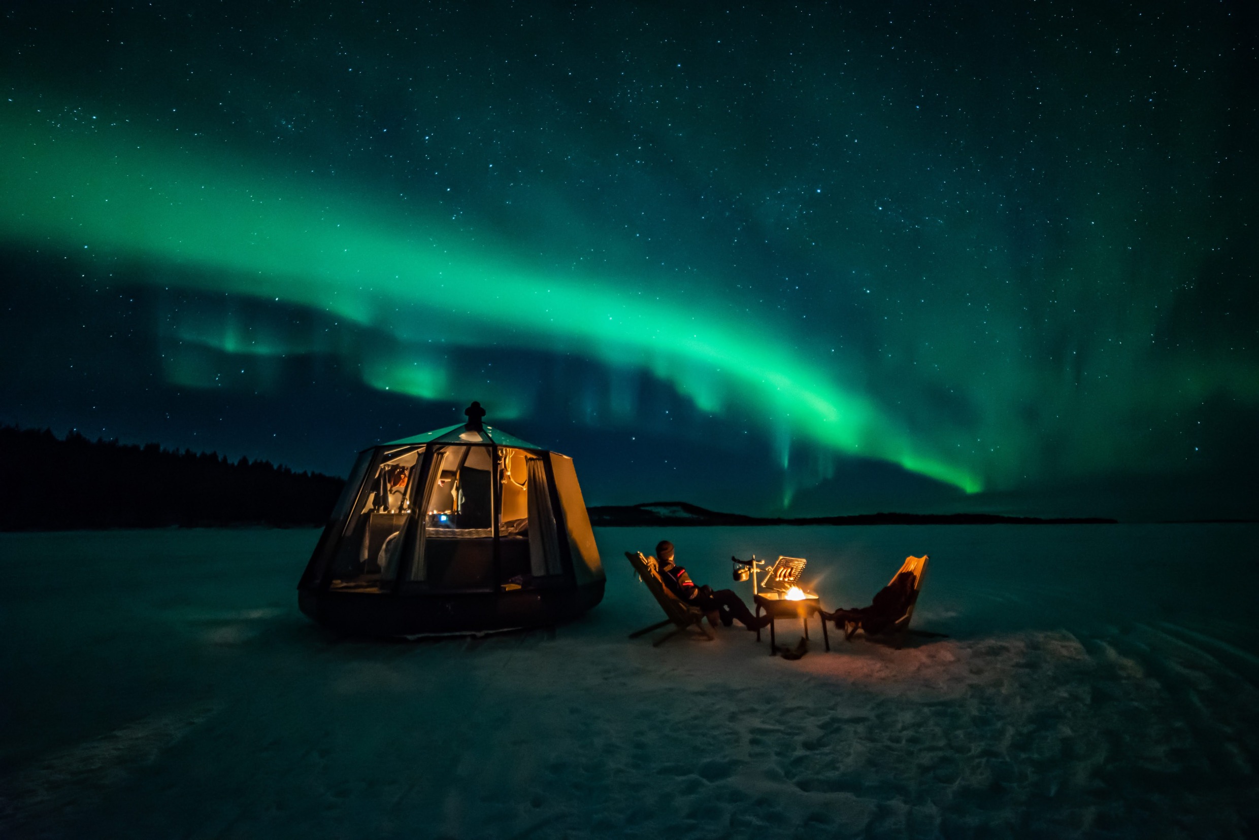 Floating glass cabin on a frozen river with firepit and Northern Lights glowing above in Jokkmokk.