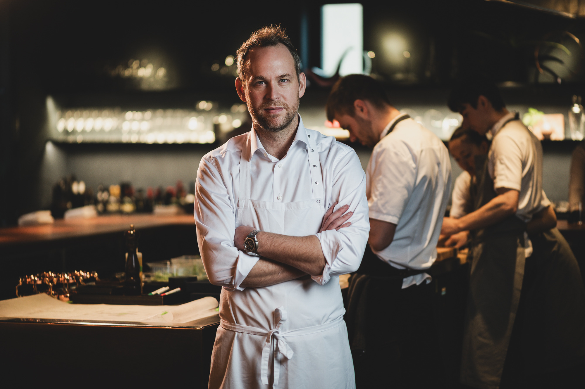 Das Bild zeigt Chefkoch Björn Frantzen in einer weißen Schürze, der in einer Restaurantküche steht, während andere Köche im Hintergrund arbeiten.
