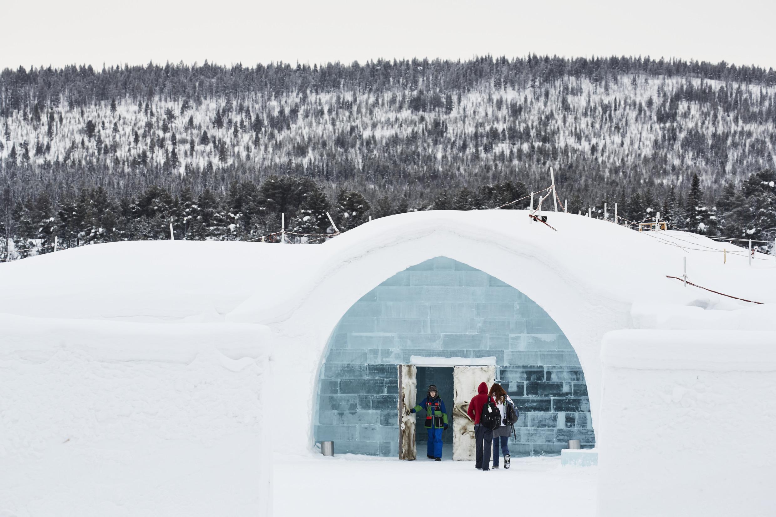 In a forest landscape, people enter the Icehotel, a building made of ice and packed snow, through a door made of ice, reindeer pelt and antlers.