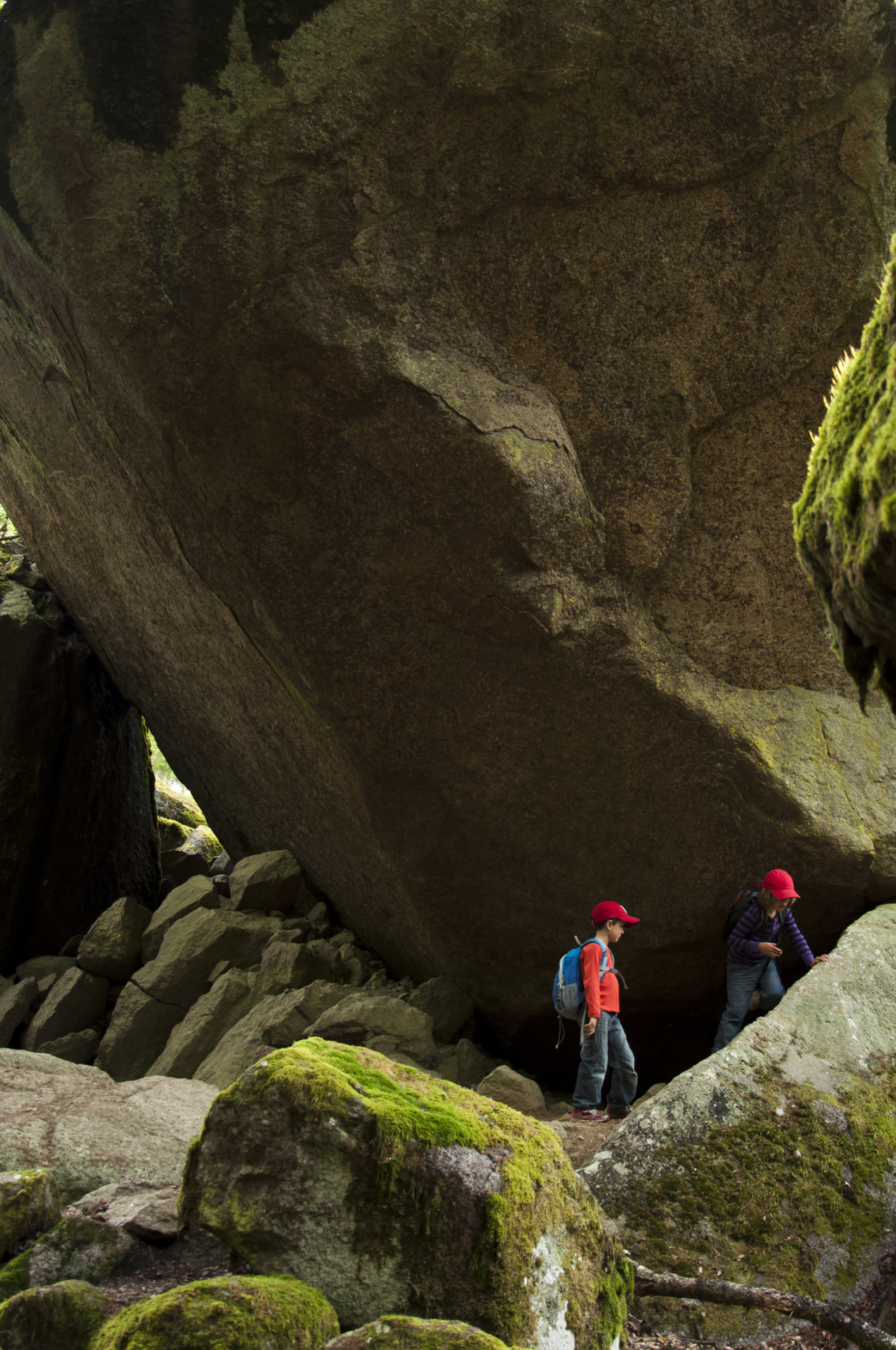 Two children wearing red caps and backpacks walking among moss-covered boulders in Tiveden National Park, with a massive rock formation towering above them.