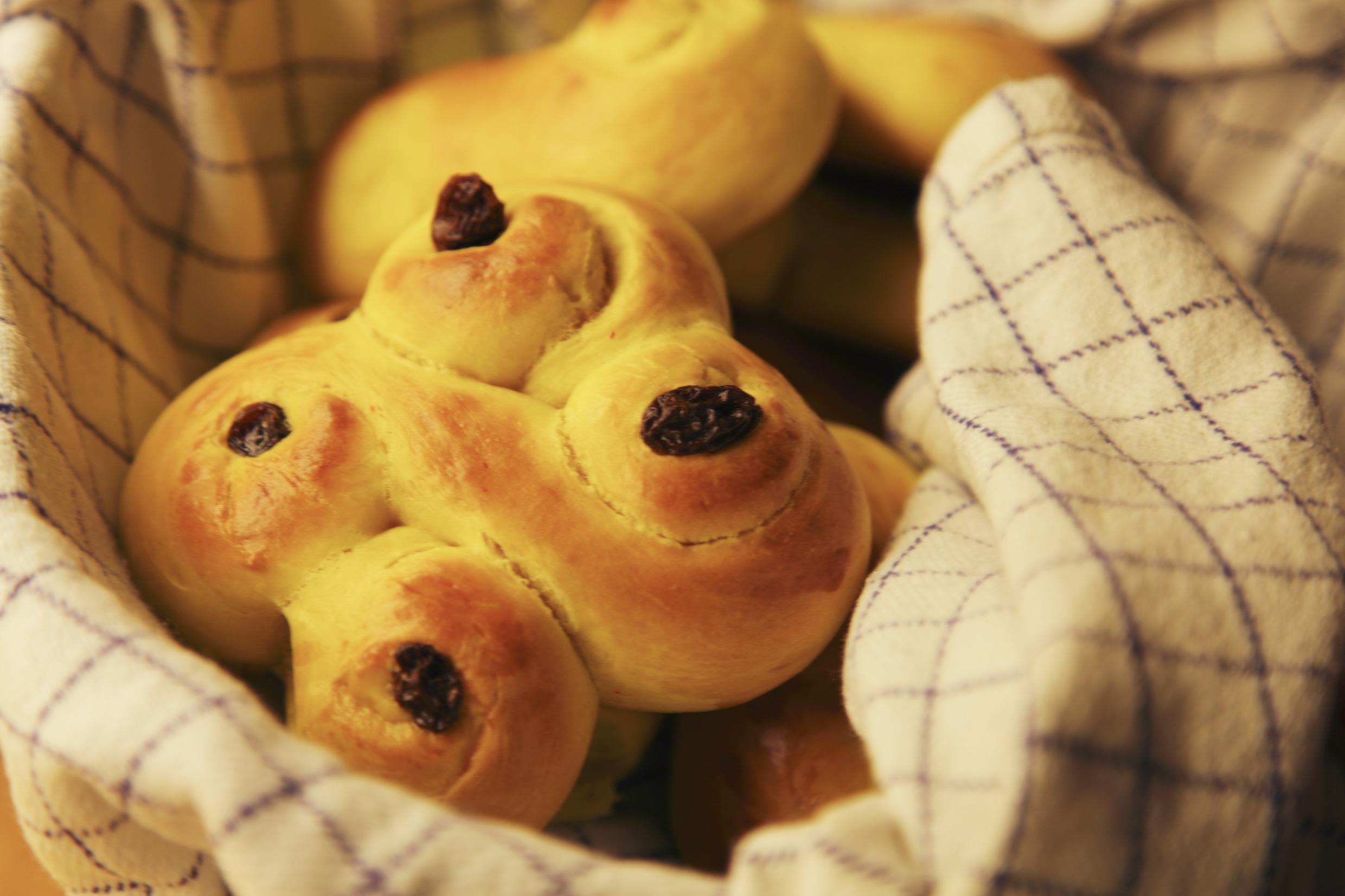 Een mand met gele saffraanbroodjes met daarop rozijnen. De broodjes zijn een vierkant dat bestaat uit twee met elkaar verweven achten.