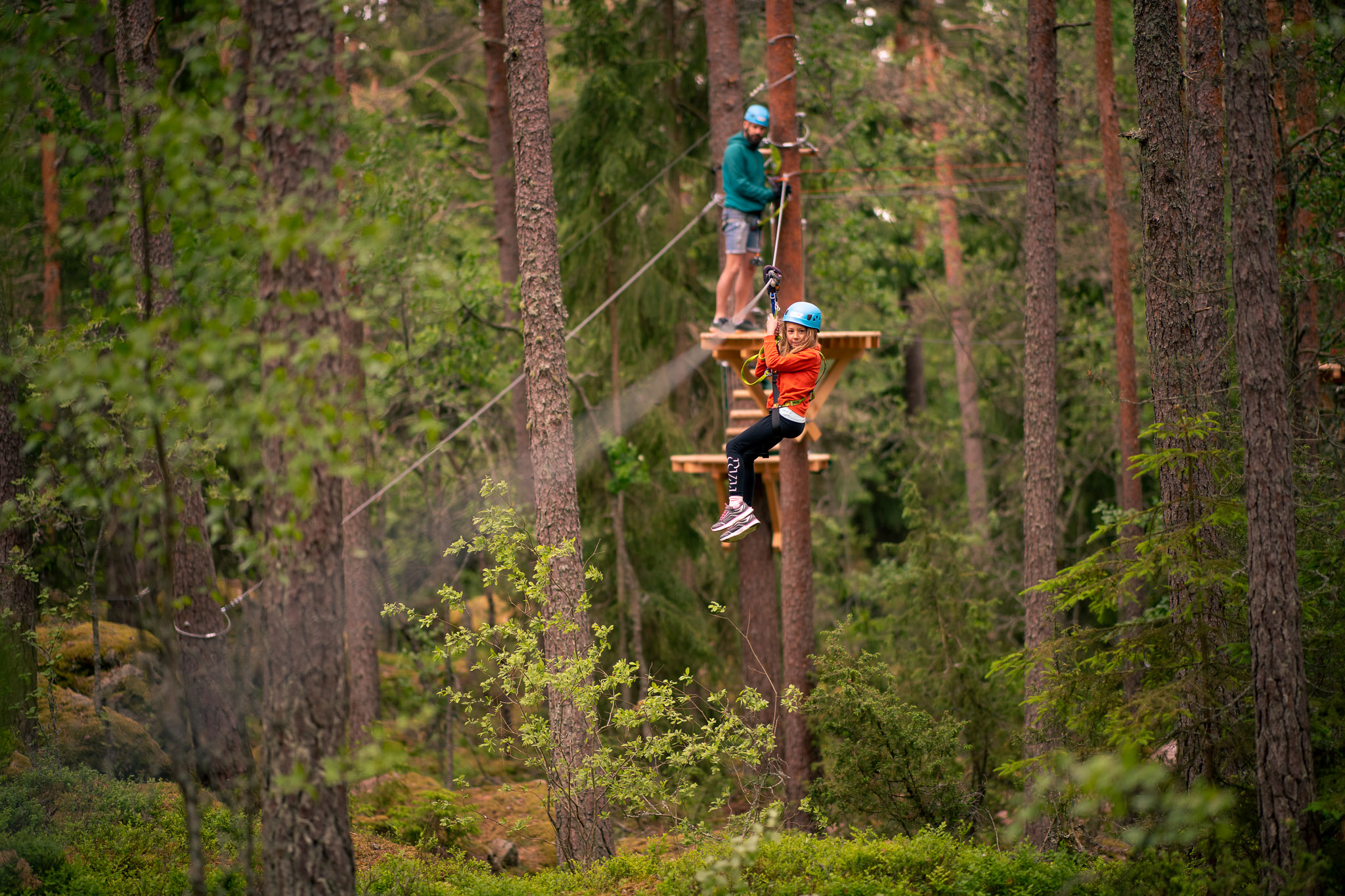 Ein Kind saust mit Helm und Sicherheitsgurt durch einen Waldabenteuerpark, während jemand anderes auf einer Plattform im Hintergrund wartet.