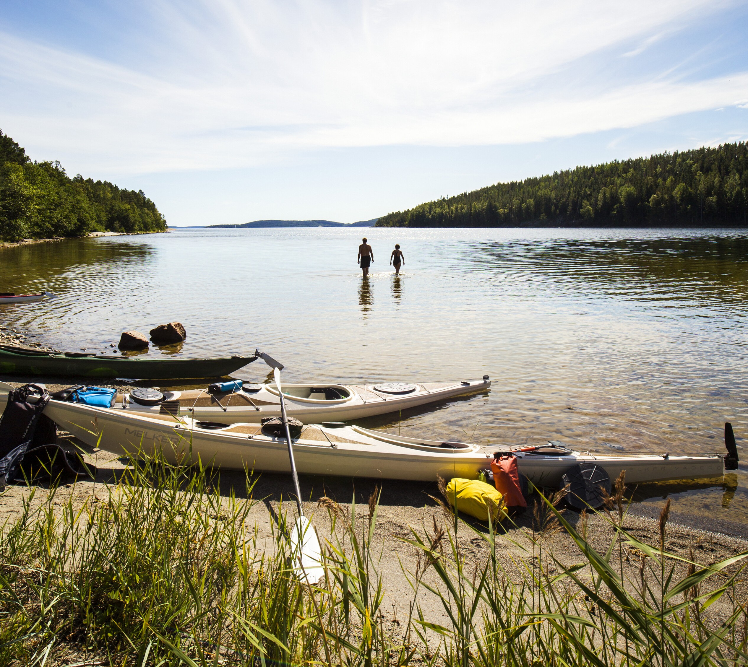 Three kayaks lay on a beach next to the water on a sunny summer day. Two people are about to take a swim. One person stands by the kayak.