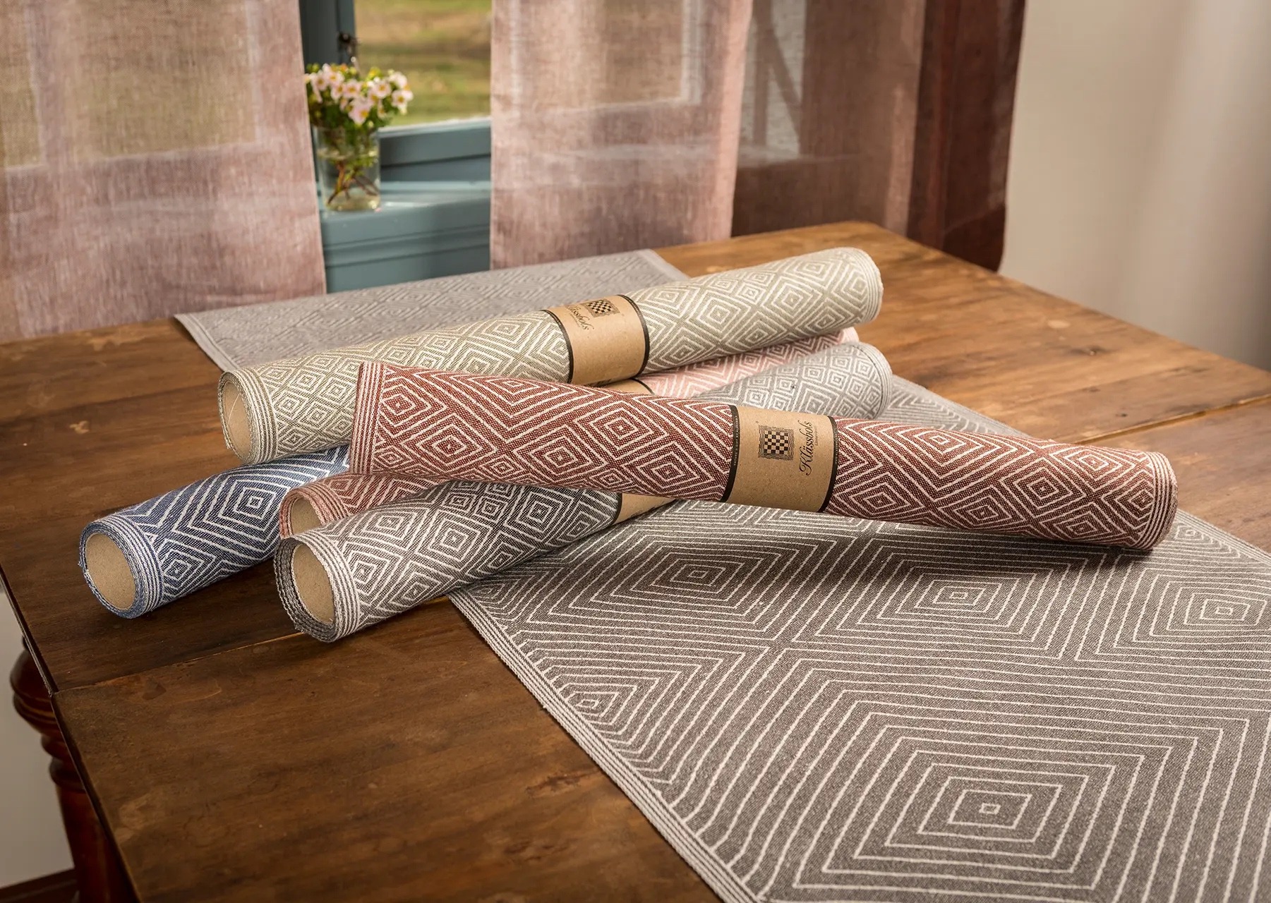 Vier Rollen Leinentischdecken auf einem Holztisch. Im Hintergrund hängen Leinenvorhänge im Fenster.