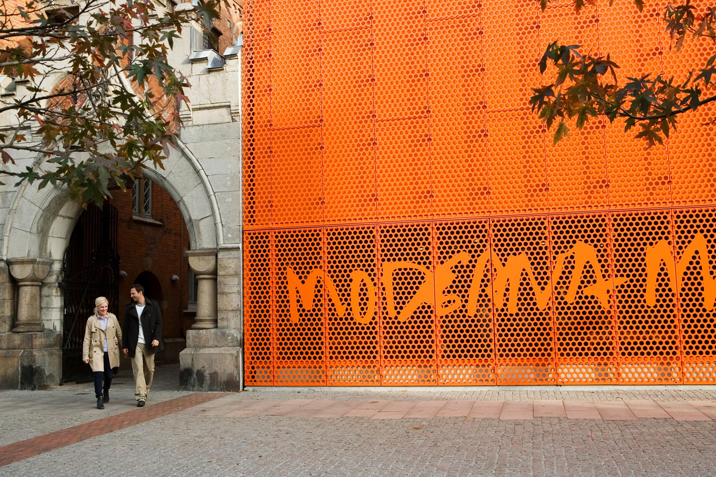 A man and a woman are walking outside and past the orange sign of the Moderna Museet in Malmö.